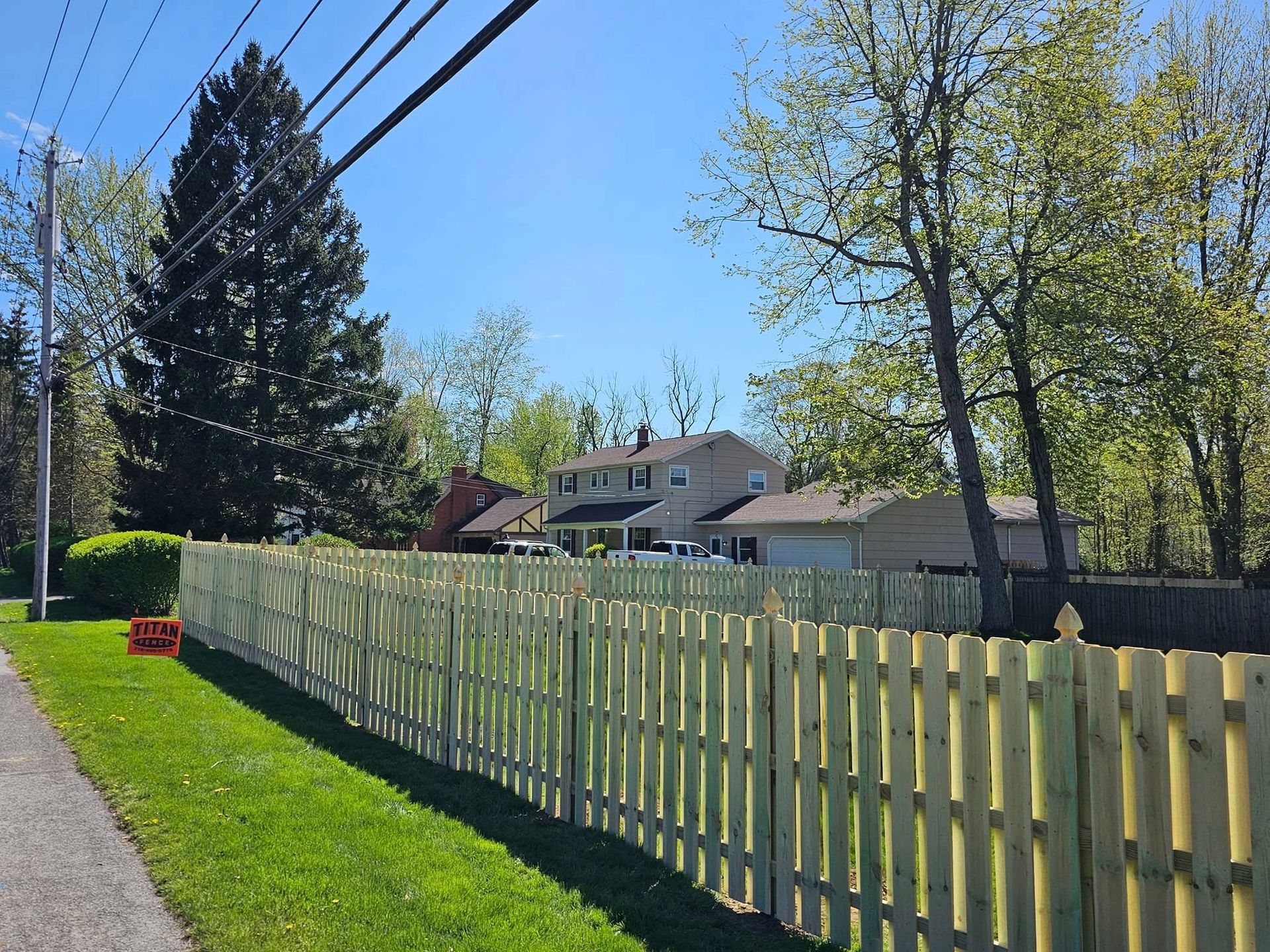 Sunlit suburban street with a long white fence, green lawn, trees, and houses under a clear blue sky