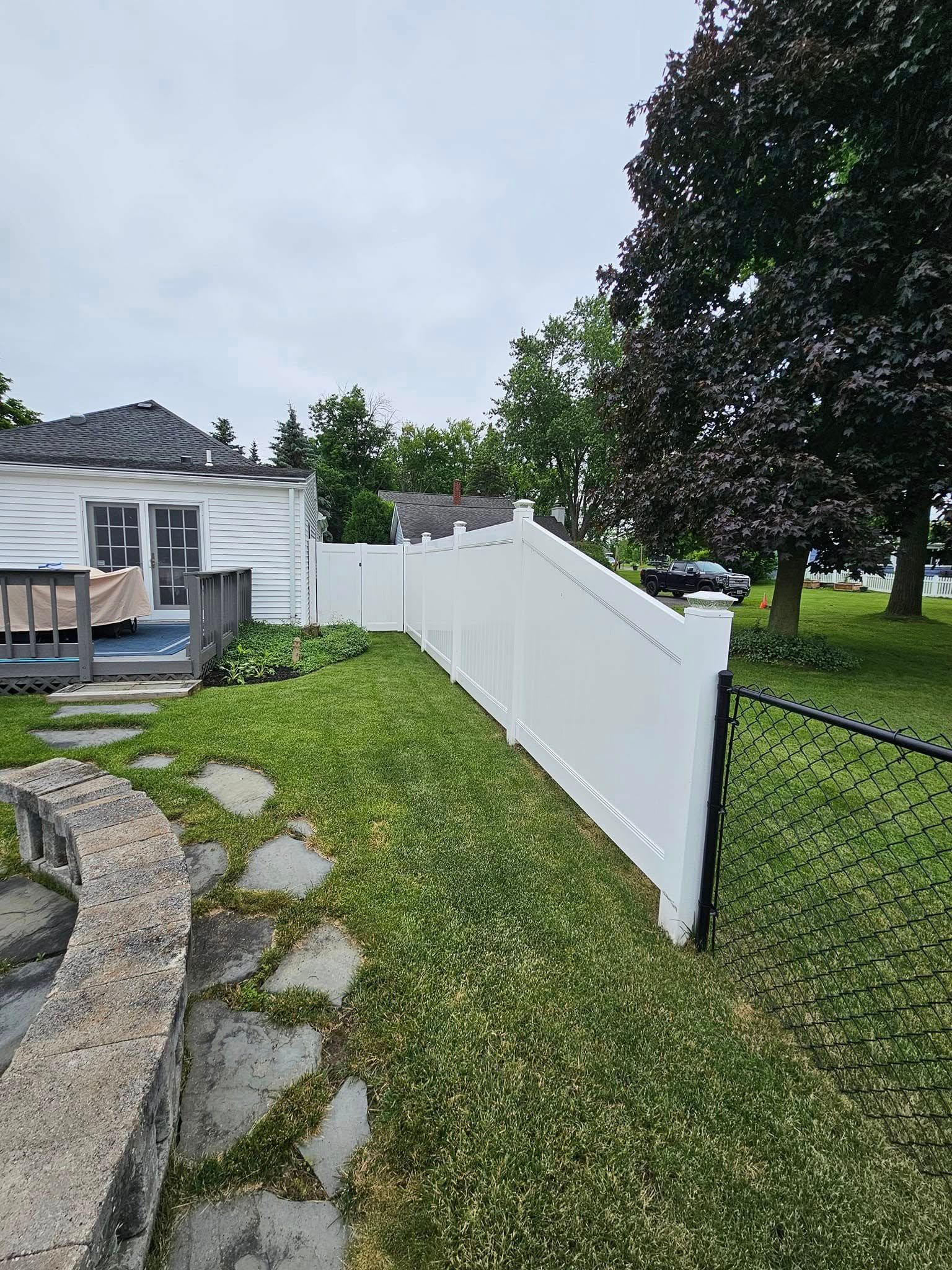White vinyl fence along a grassy backyard beside a house, stone path, and large tree under a cloudy sky