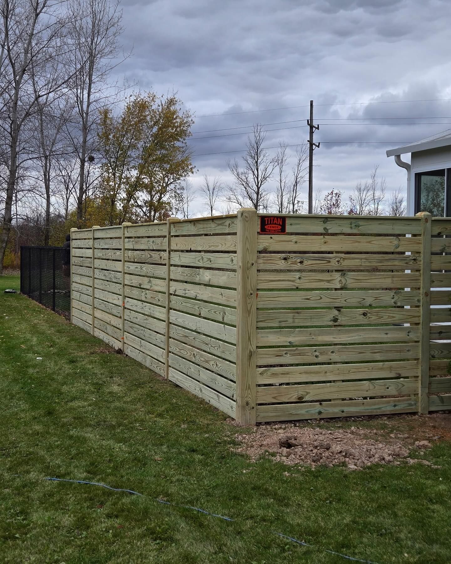 Wooden slat fence along a grassy yard under cloudy skies, with trees and a house corner nearby.