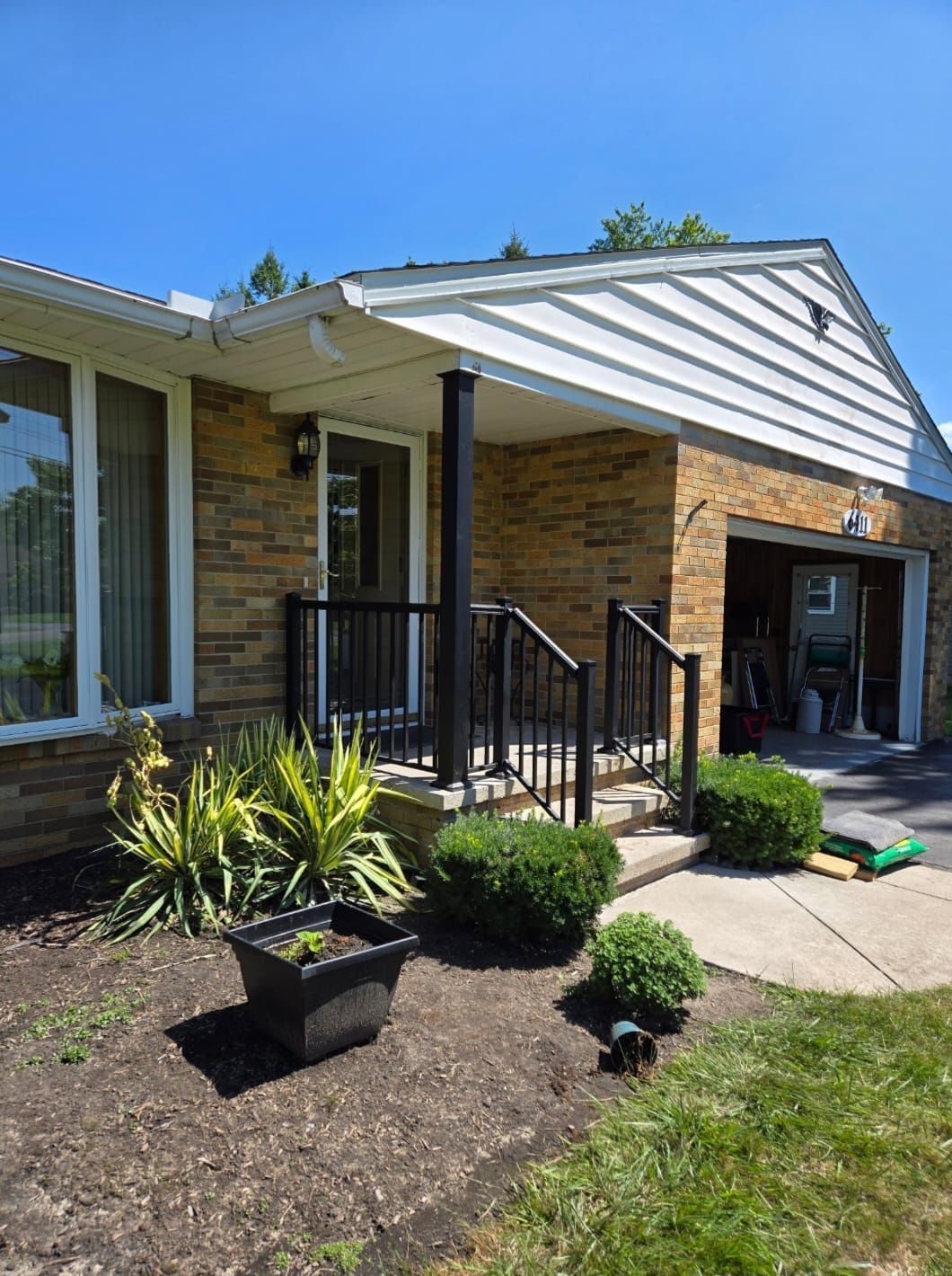 Front porch of a brick house with black railing, potted plants, and an open garage in bright sunlight