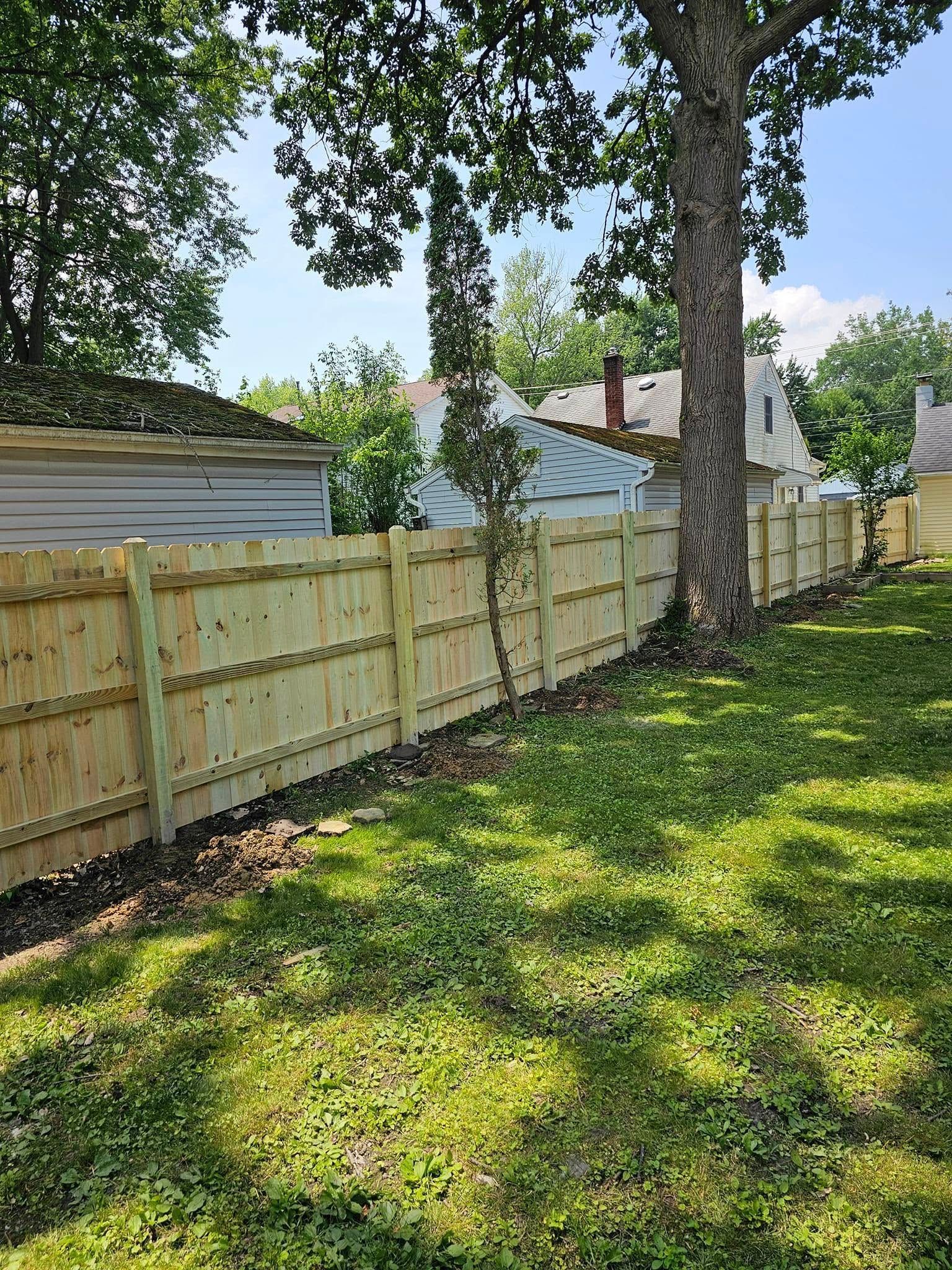Backyard with wooden fence, tall trees, and a sunlit grassy lawn beside houses