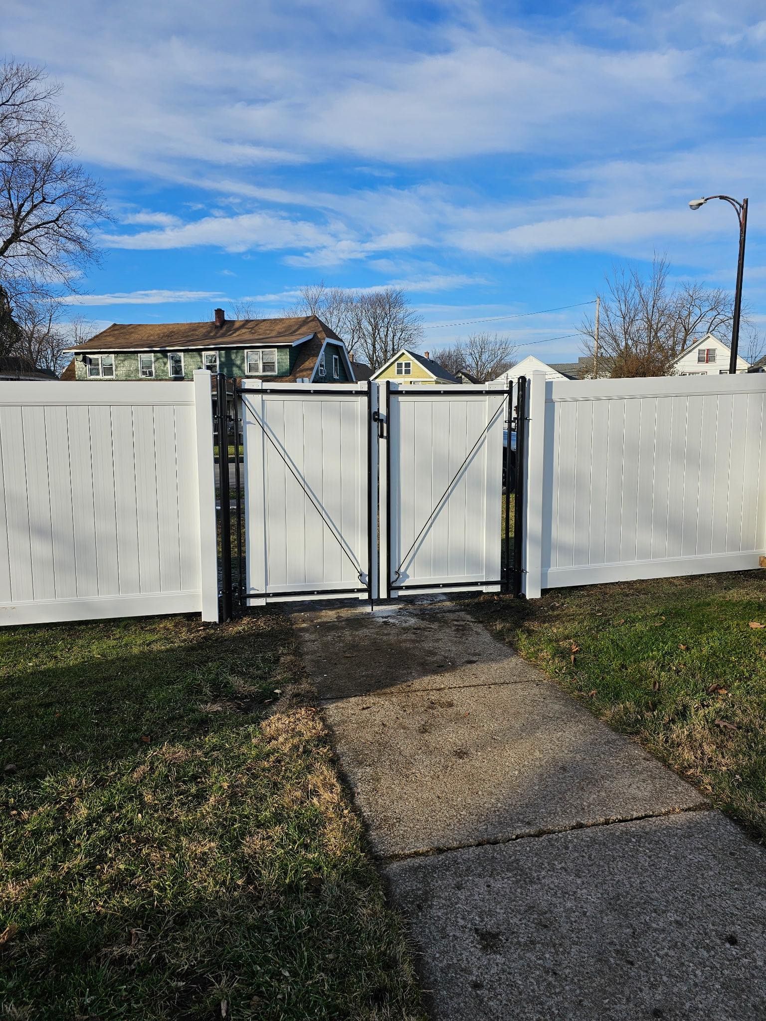 White metal gate in a fenced yard with a paved path, blue sky, and house in the background