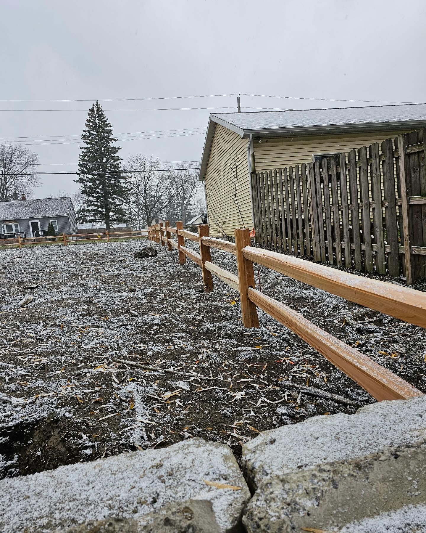 Snow-dusted farmyard with a wooden fence, shed, and trees under a gray winter sky
