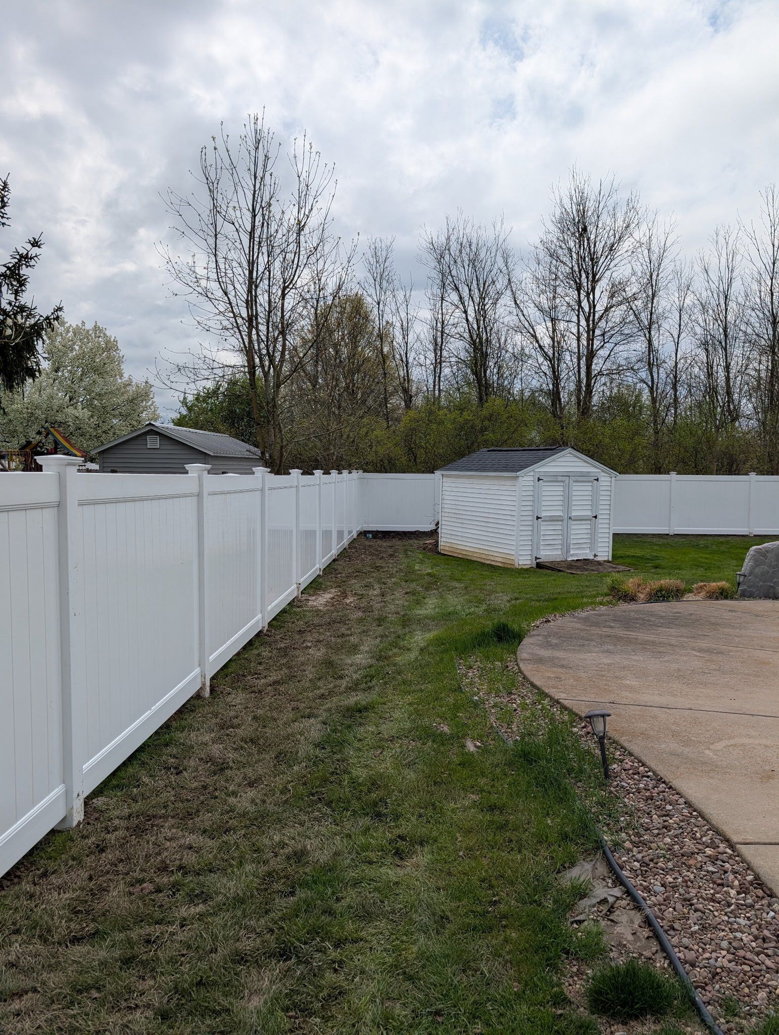 White vinyl fence lines a grassy side yard beside a gravel driveway under a cloudy sky.