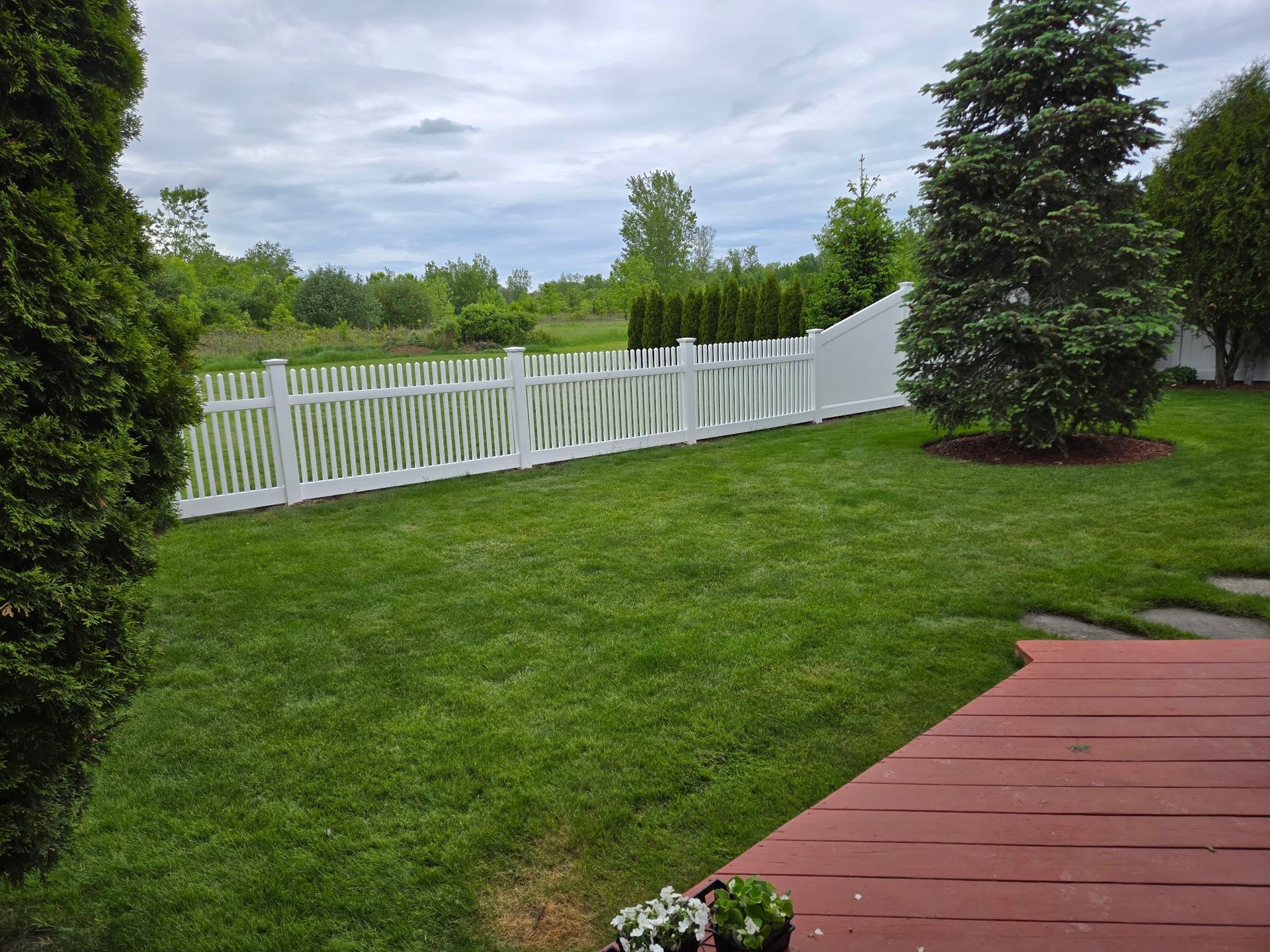 Backyard with green lawn, white picket fence, and red wooden deck under cloudy sky