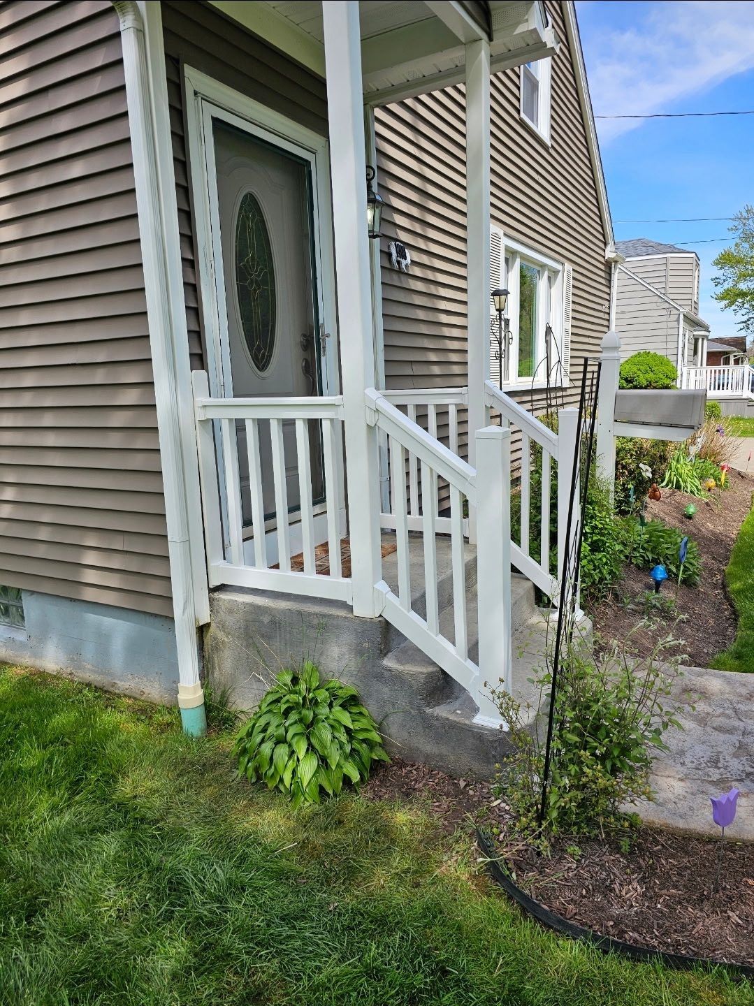 Front porch of a tan house with white railing, steps, and a small garden beside the walkway.