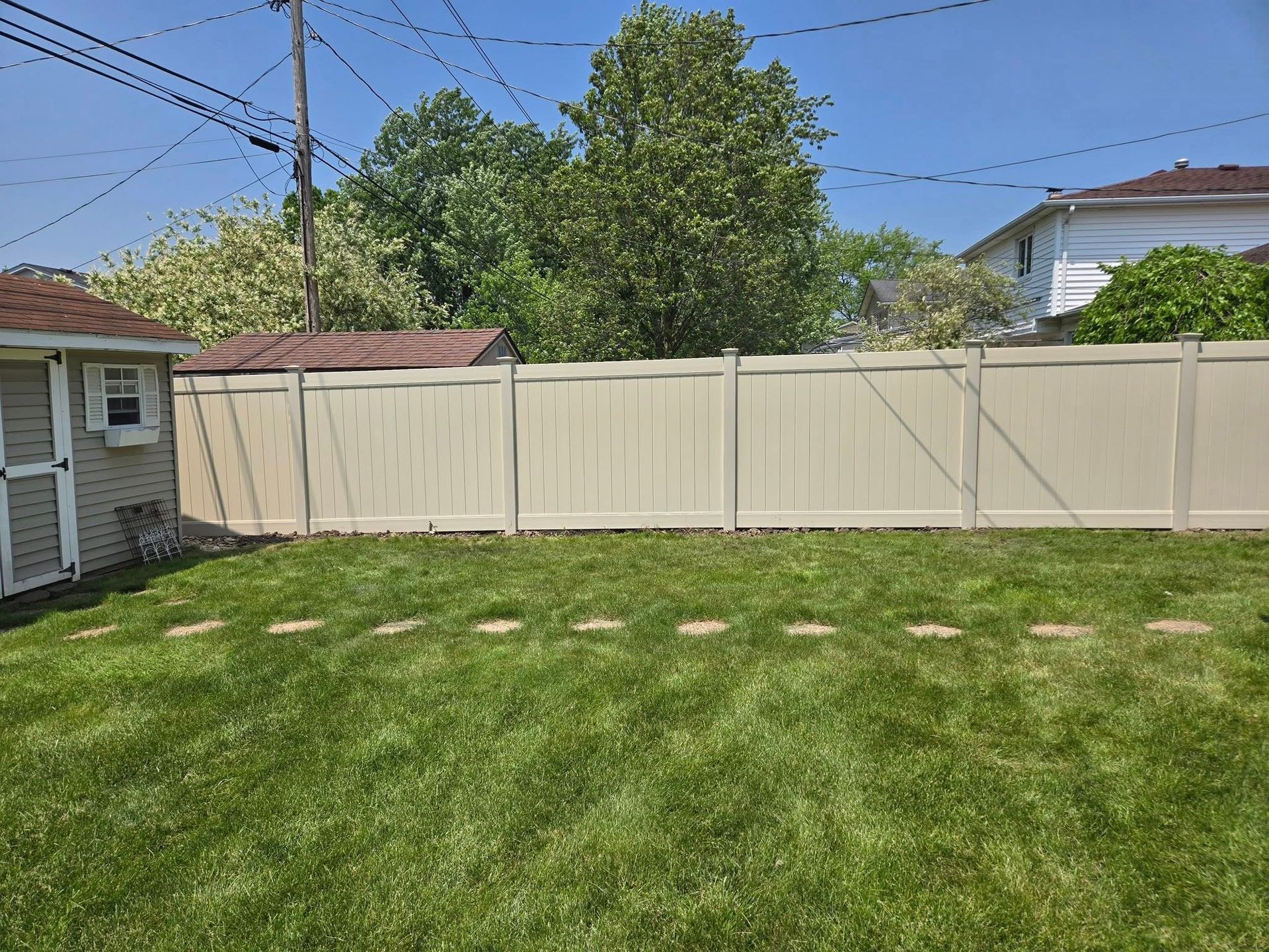 Backyard with green lawn, beige fence, trees, and a small house under a blue sky