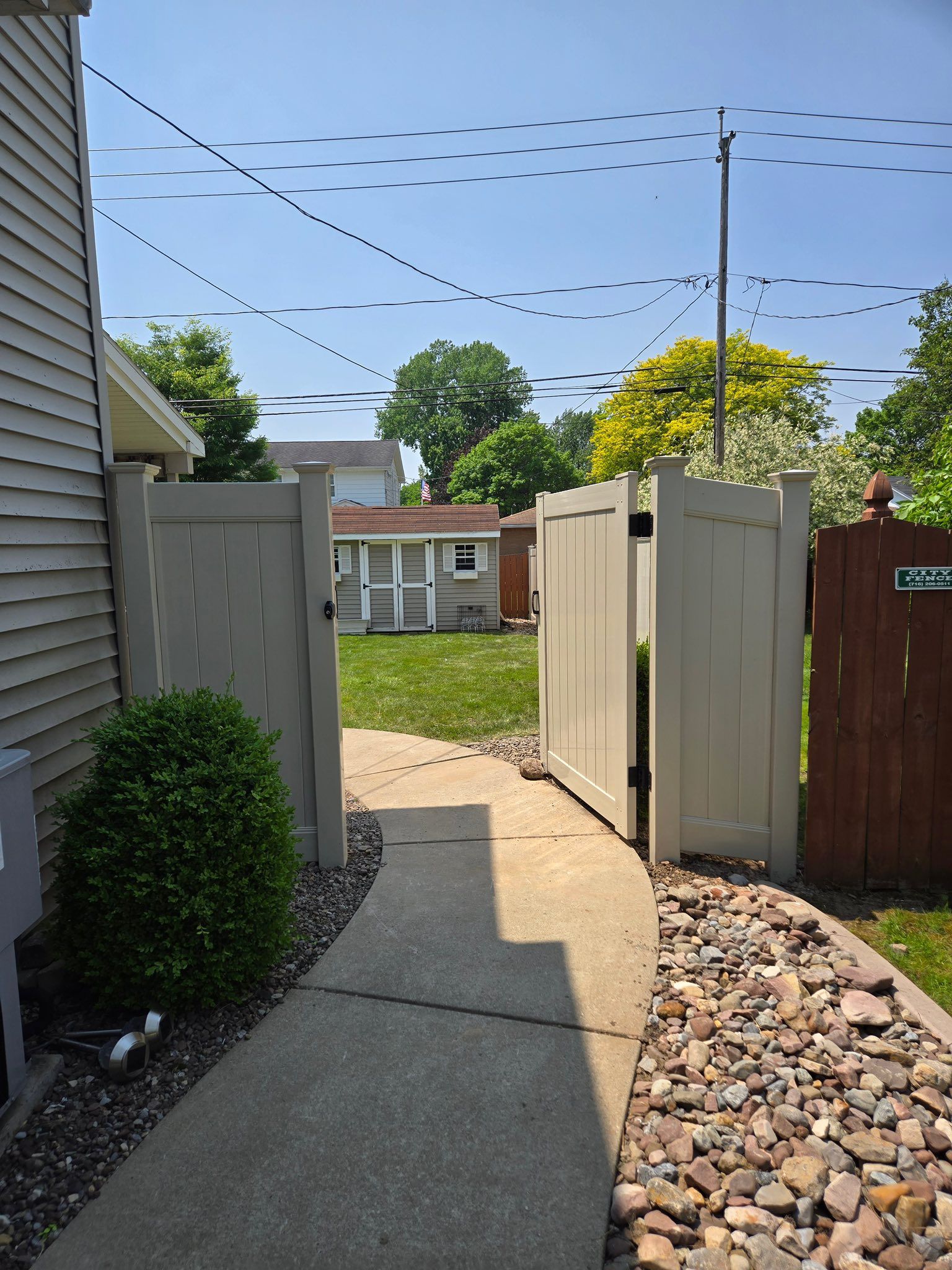 Narrow concrete path between beige fences leading to a backyard, with rocks and a clear blue sky.