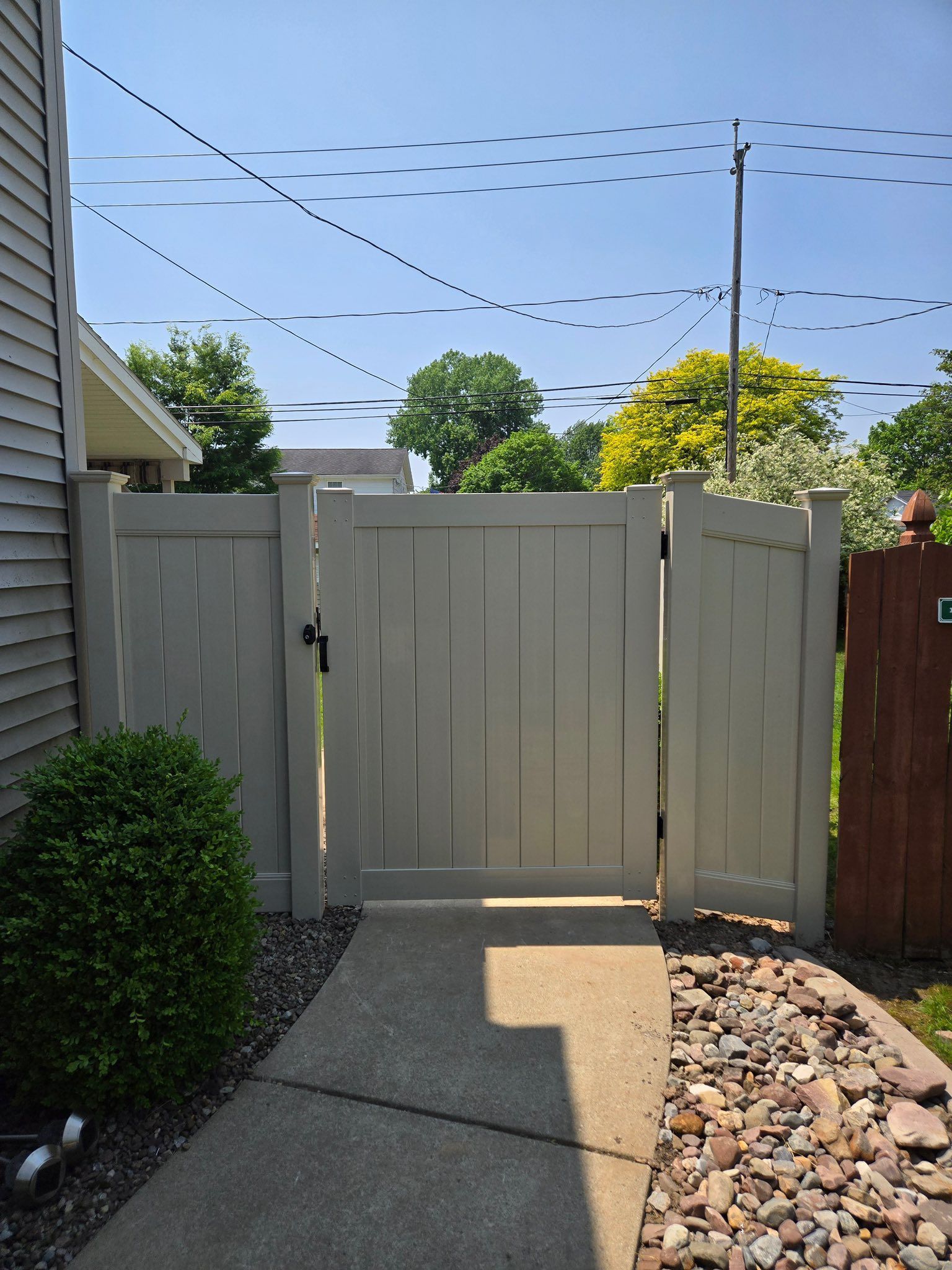 White wooden backyard gate between a gray house and fence, with rocks and a concrete path in bright sunlight.