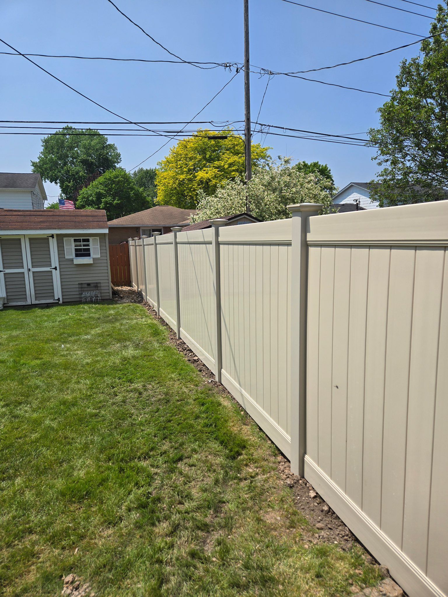 Backyard with beige vinyl fence, grassy lawn, shed, trees, and blue sky with power lines