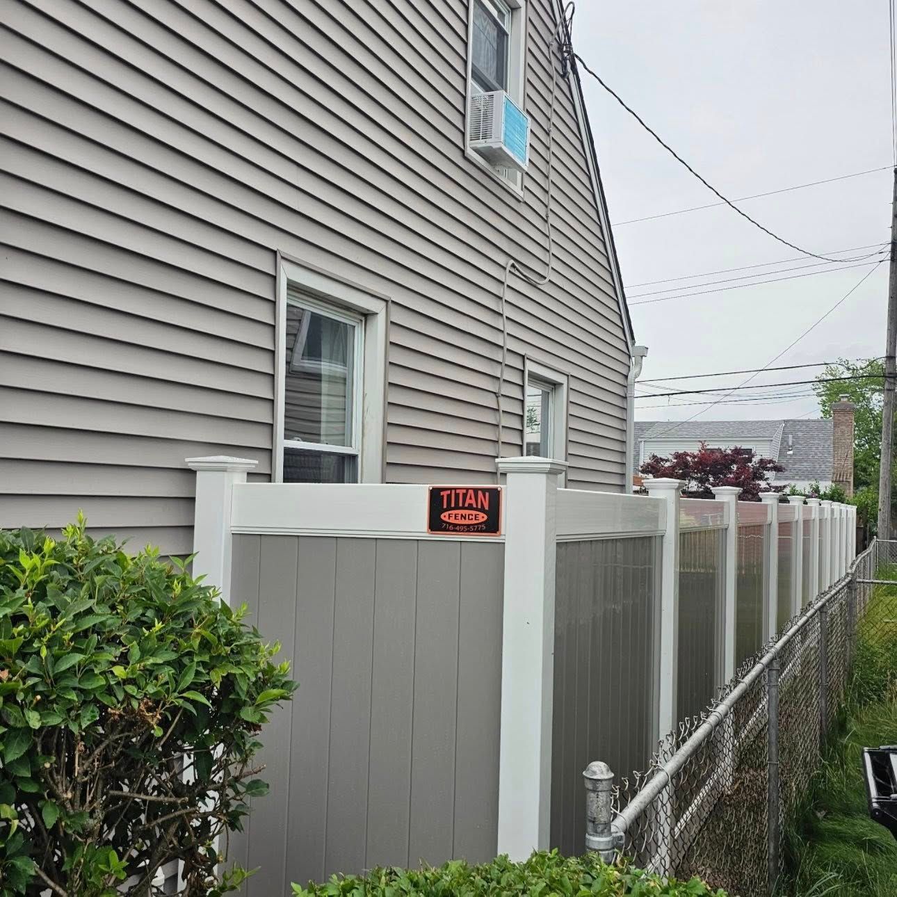 Side of a gray house with white fence, hedge, and narrow walkway beside a chain-link fence.