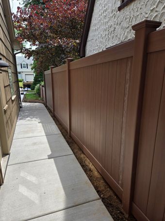 Narrow concrete walkway beside a brown wooden fence and building, with trees and houses ahead.