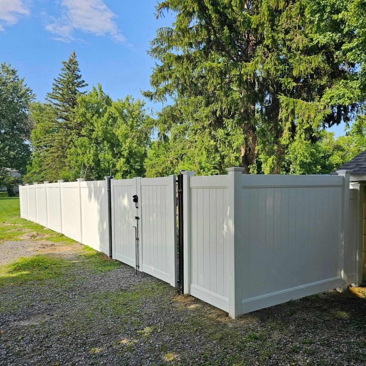 White vinyl fence and gate along a gravel yard under sunny blue sky, with tall green trees behind.