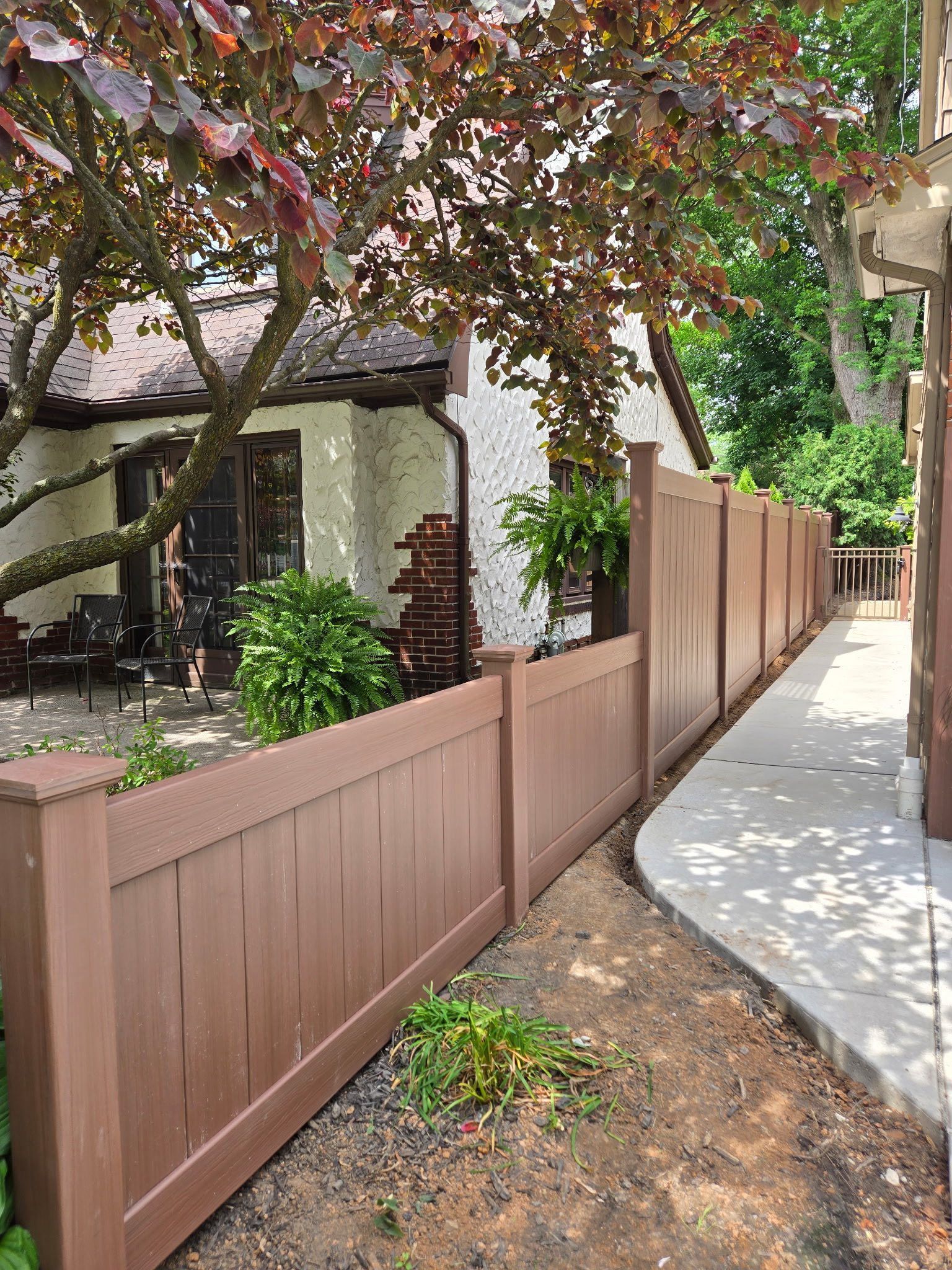 Brown wooden fence along a sidewalk beside a leafy residential yard and house