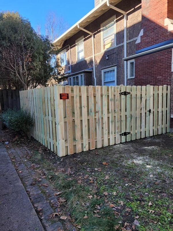 Wooden fenced enclosure beside a brick apartment building, with a red warning sign near a sidewalk.
