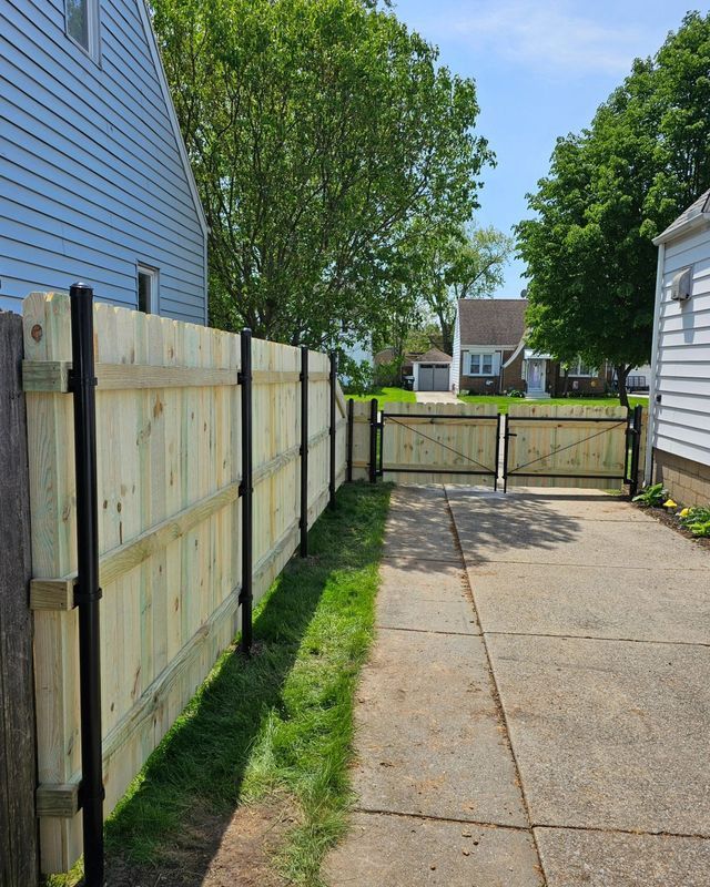 Concrete driveway beside a wooden fence, leading to a closed gate and houses beyond on a sunny day
