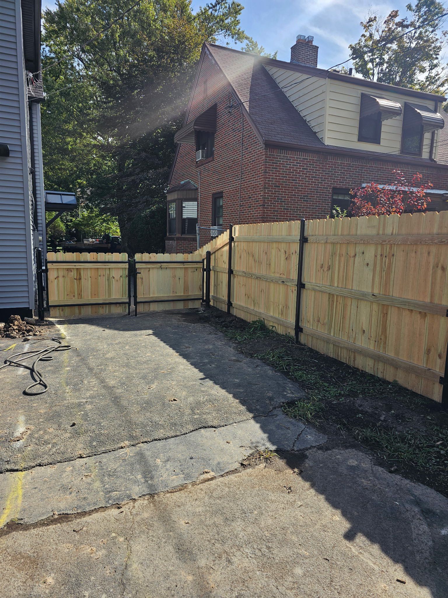 Backyard driveway beside a wooden fence and gate, with a brick house in sunlight.