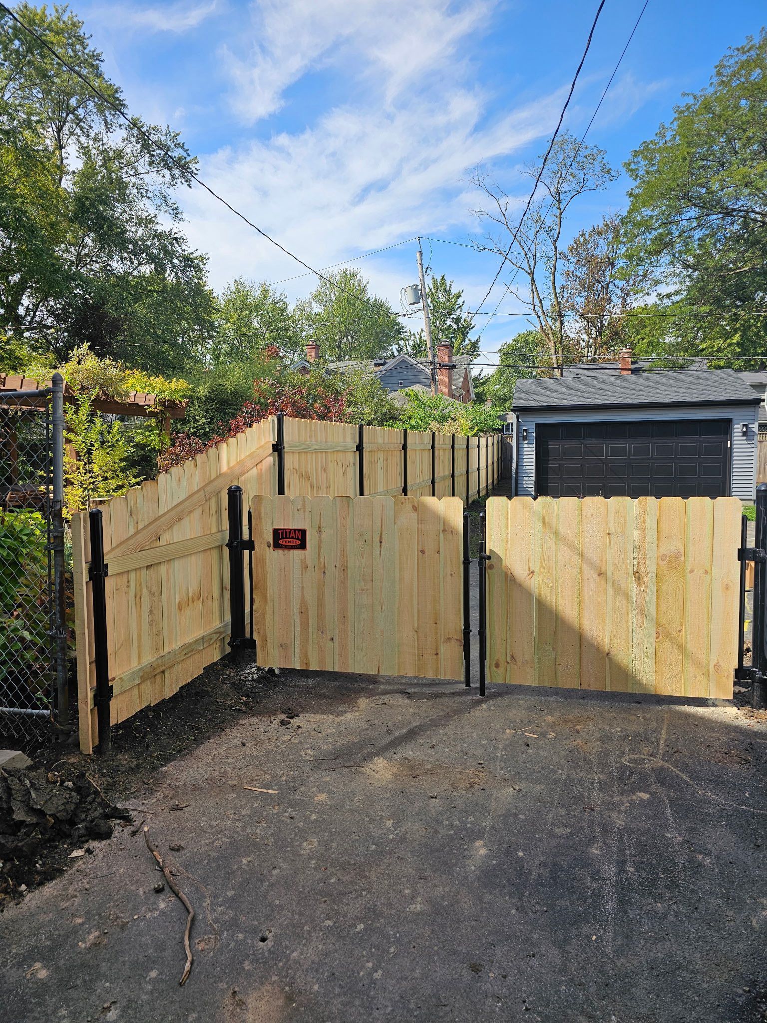 Wooden driveway gate and fence beside a garage, with gravel ground and a blue sky overhead