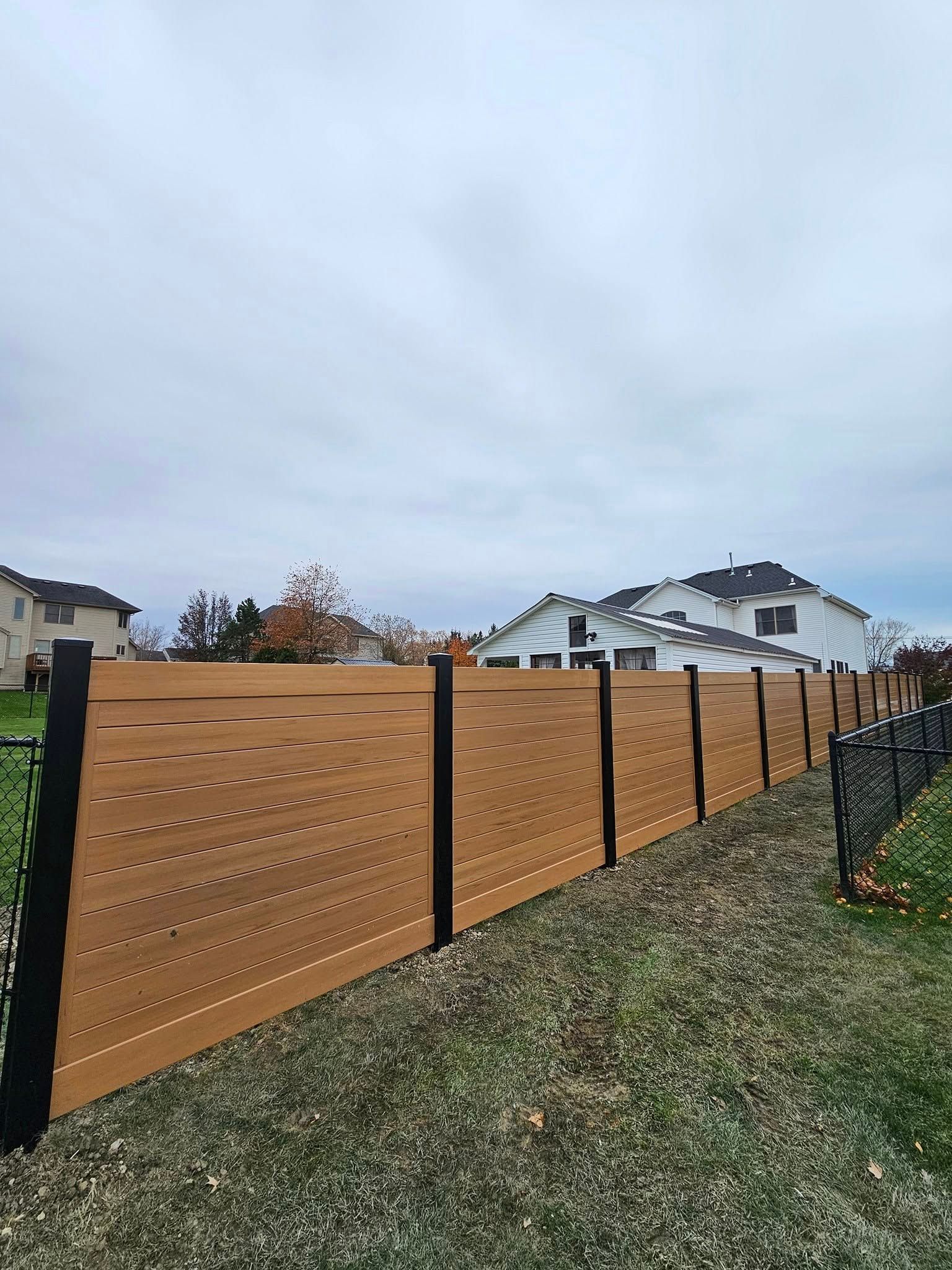 Wooden privacy fence with black posts along a grassy slope in a suburban neighborhood under an overcast sky