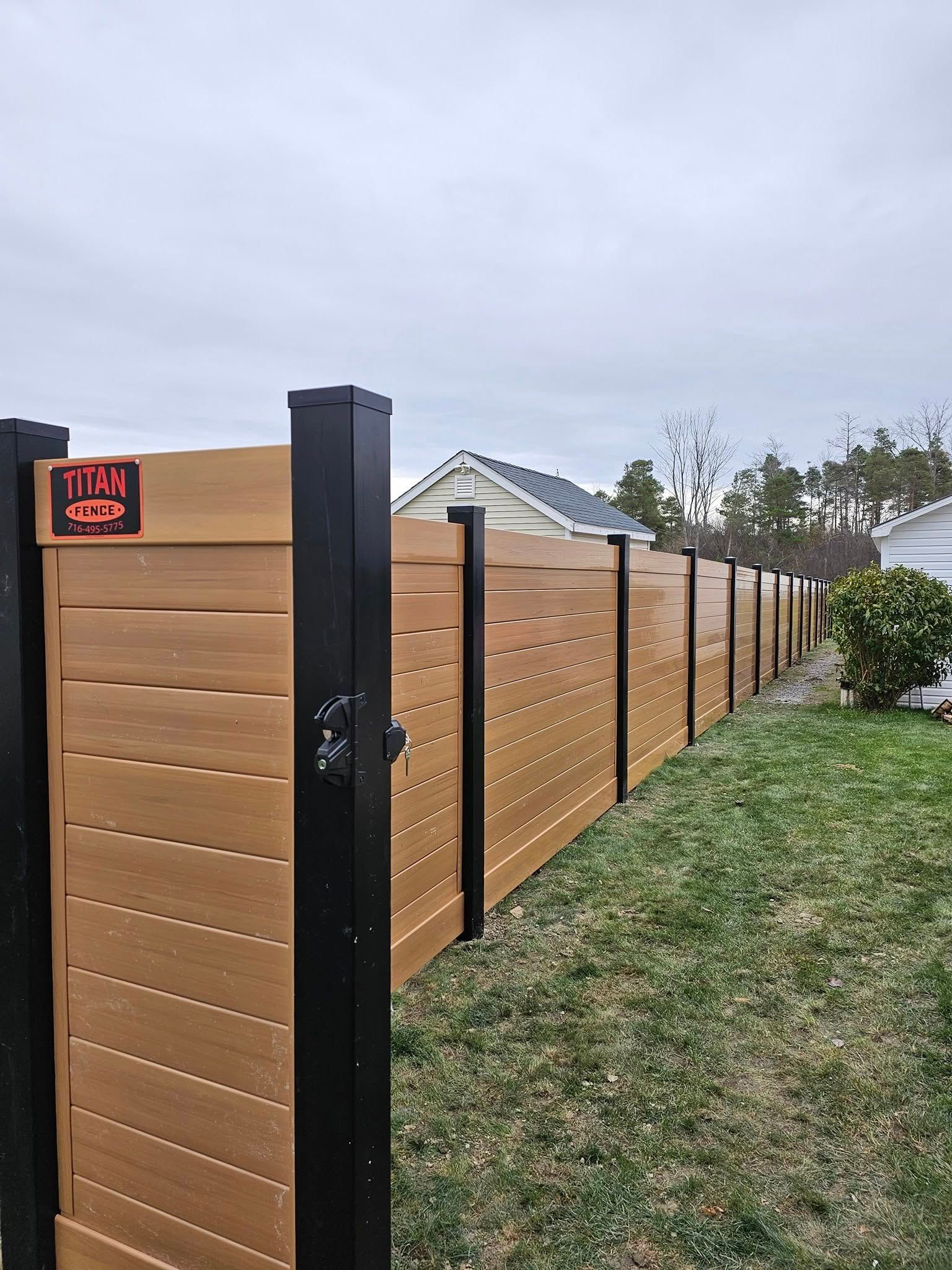 Modern wooden fence with black posts and a red alarm light along a grassy yard under cloudy sky