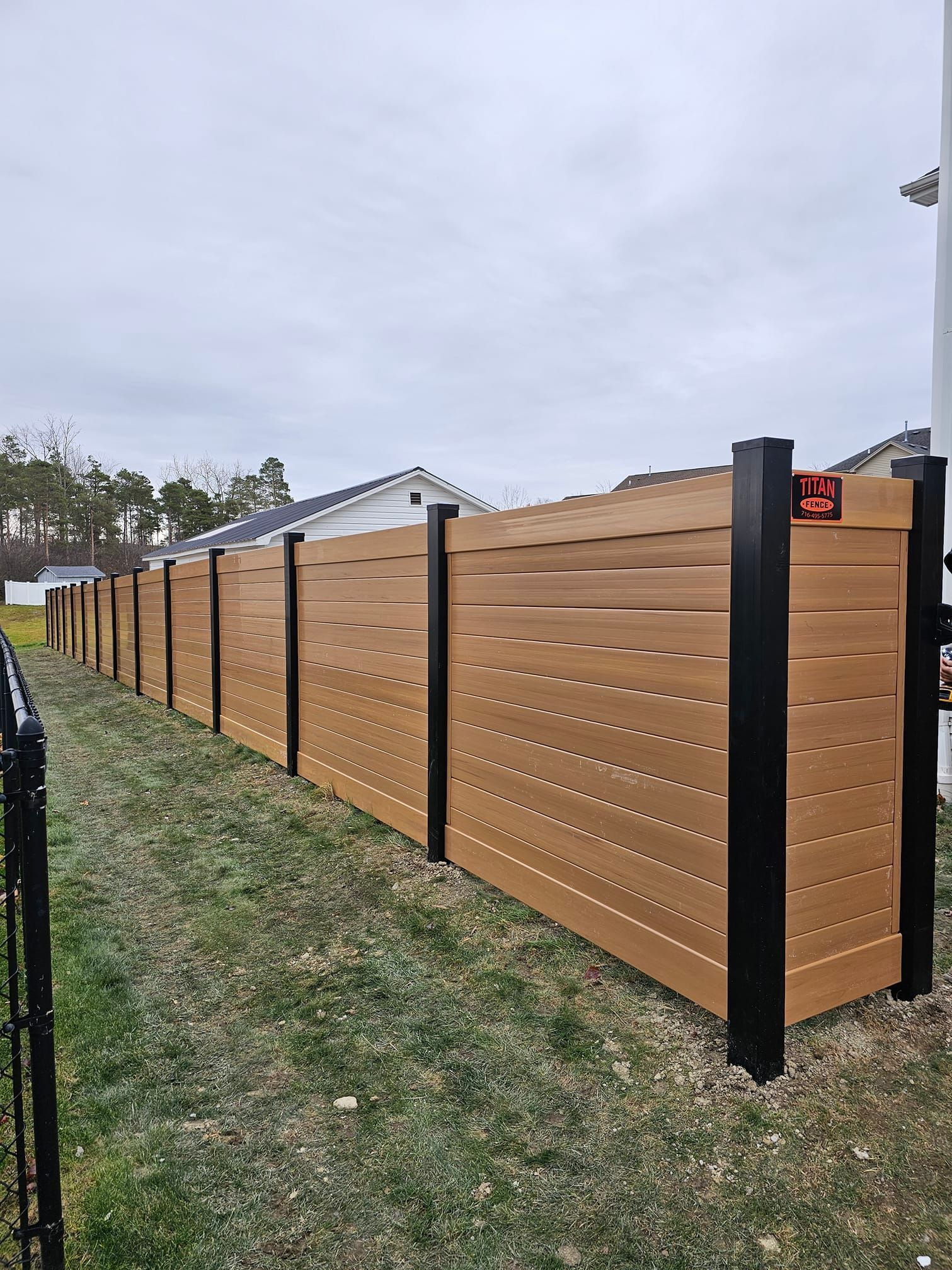 Brown slatted privacy fence with black posts running along a grassy yard under an overcast sky
