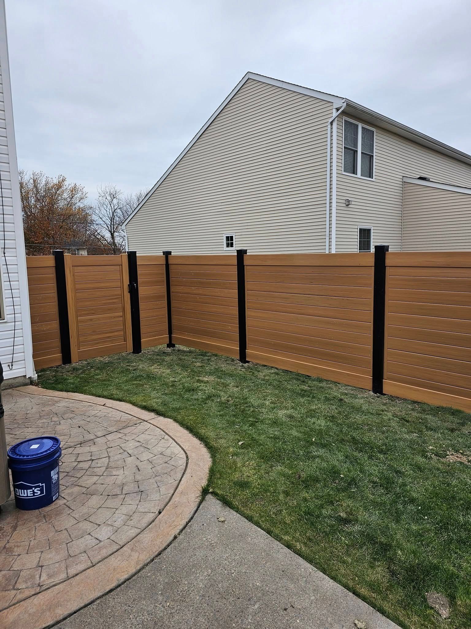 Backyard with curved wooden fence, green grass, patio stones, and a house with storm-damaged siding