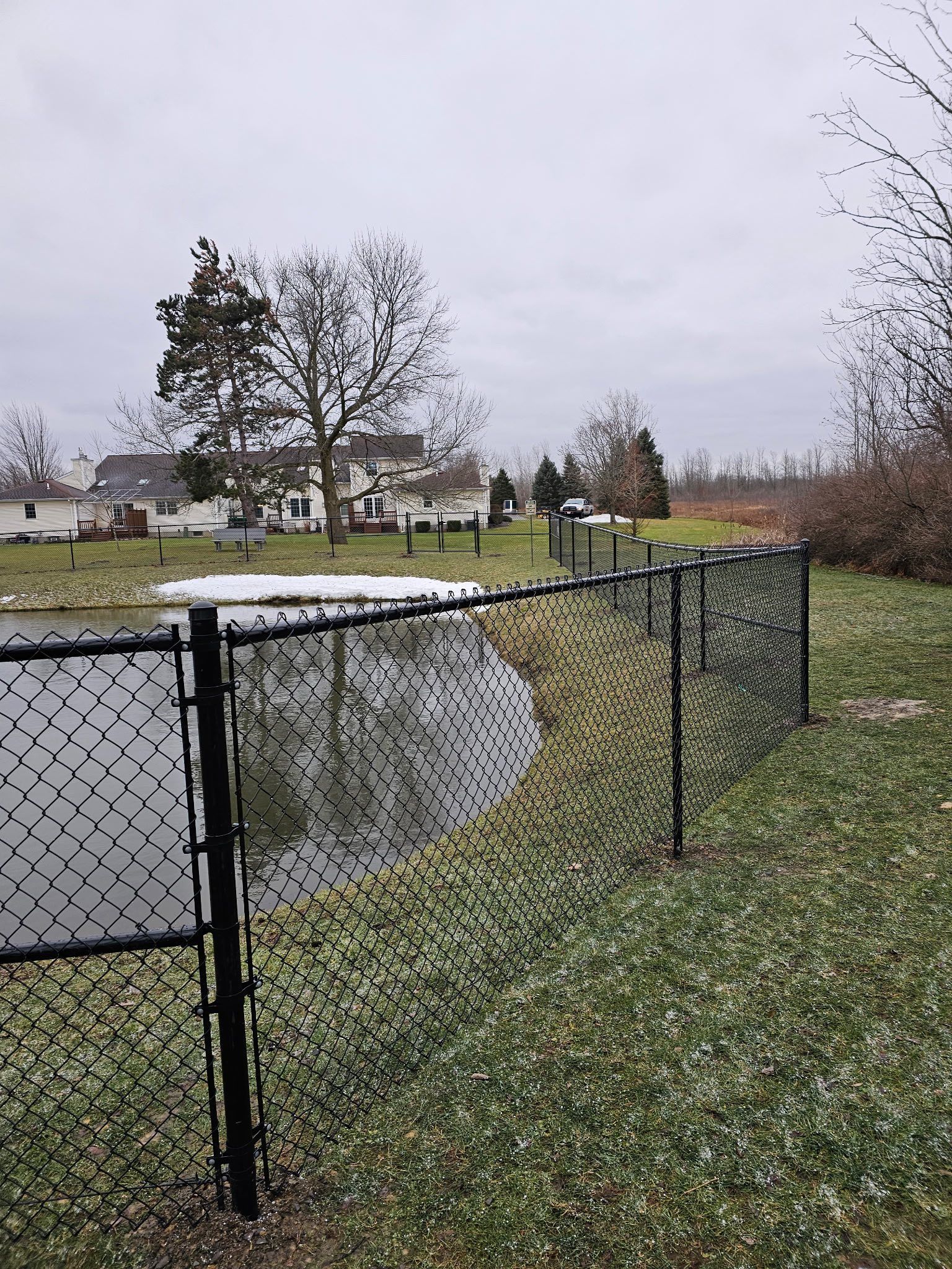 Chain-link fence beside a small pond in a grassy park on an overcast day