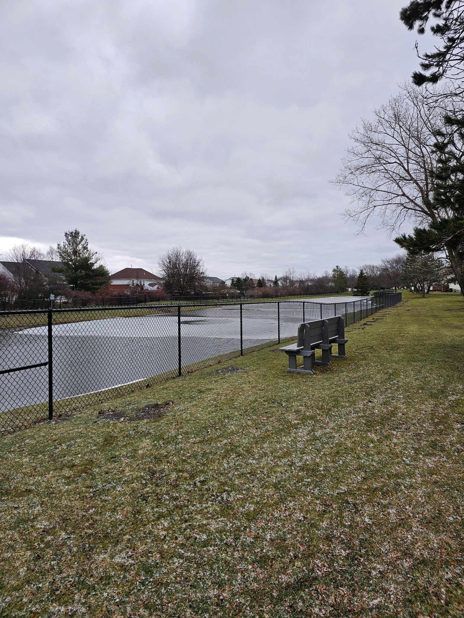Cloudy park path beside a fenced pond, with benches and light snow on the ground.