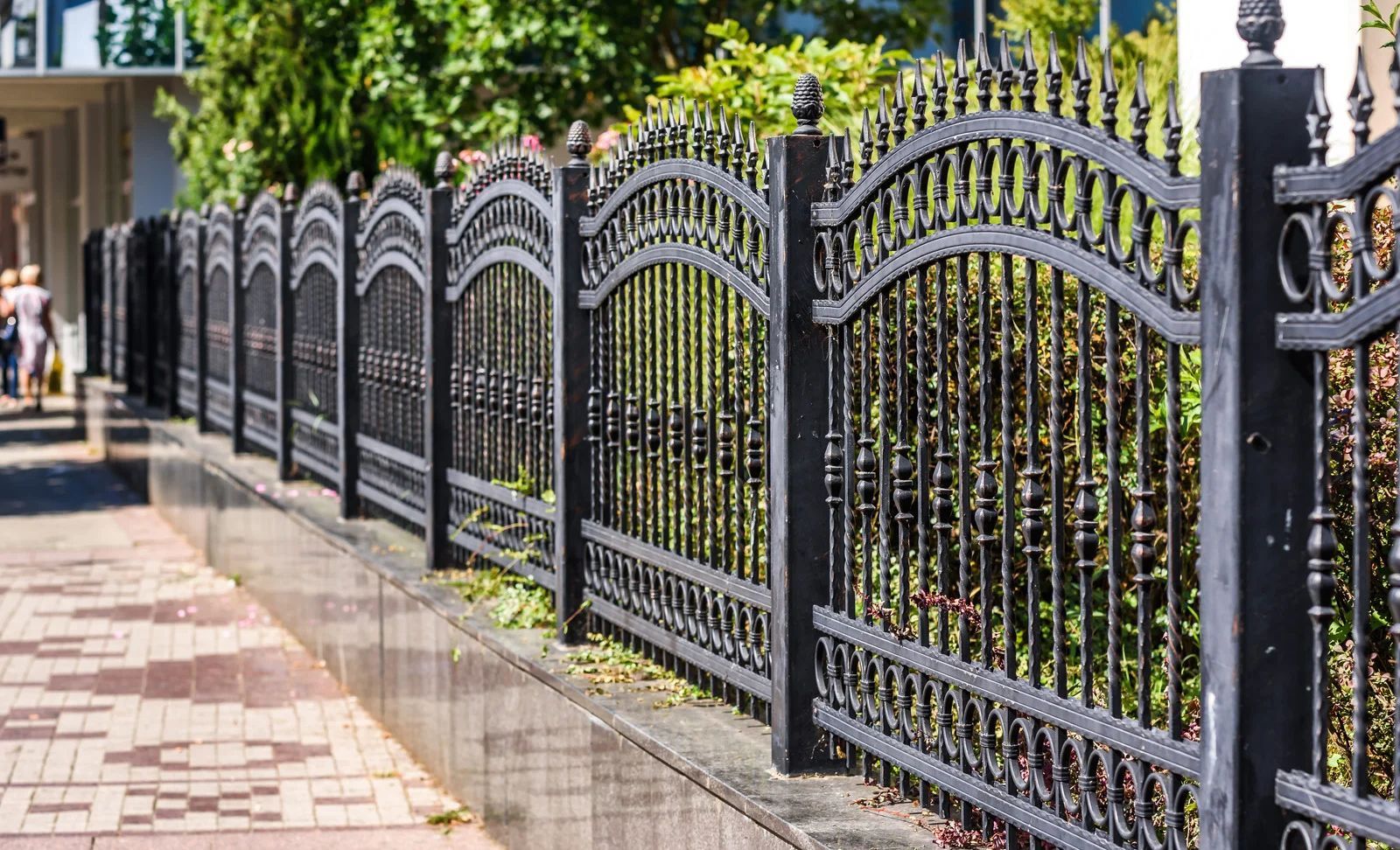 Black wrought-iron fence with arched details along a brick sidewalk beside green trees and a residential street