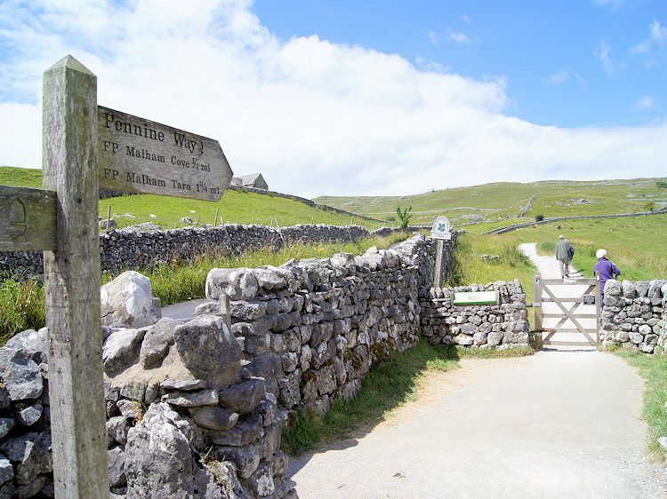 A wooden signpost stands by a stone wall near a gate on a grassy path with hikers in the distance under a blue sky.