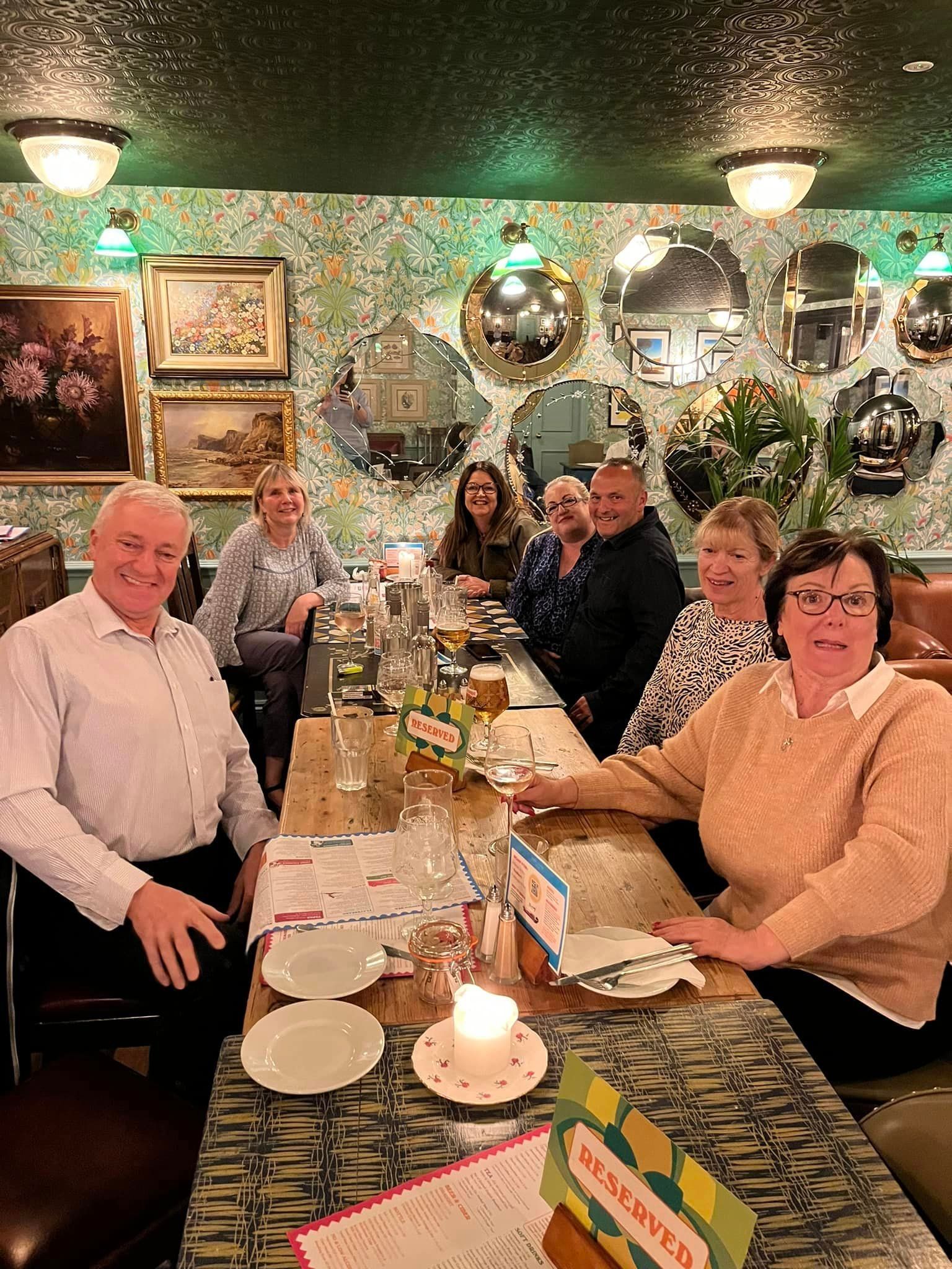 A group of eight people sitting at a table in a dimly lit, decorated restaurant with mirrors on the wall.