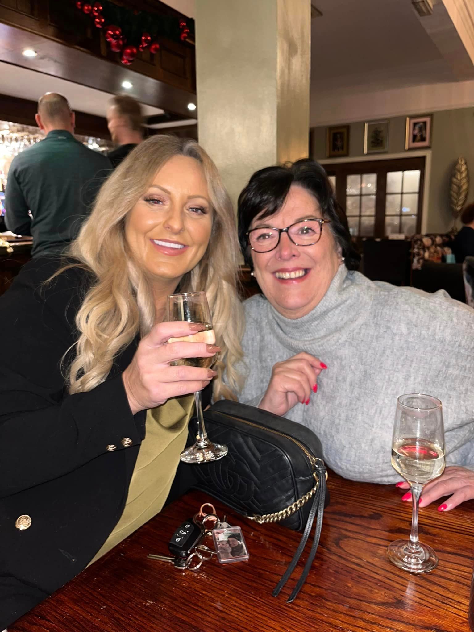 Two people smile while sitting at a wooden table in a restaurant, each holding a glass of wine.