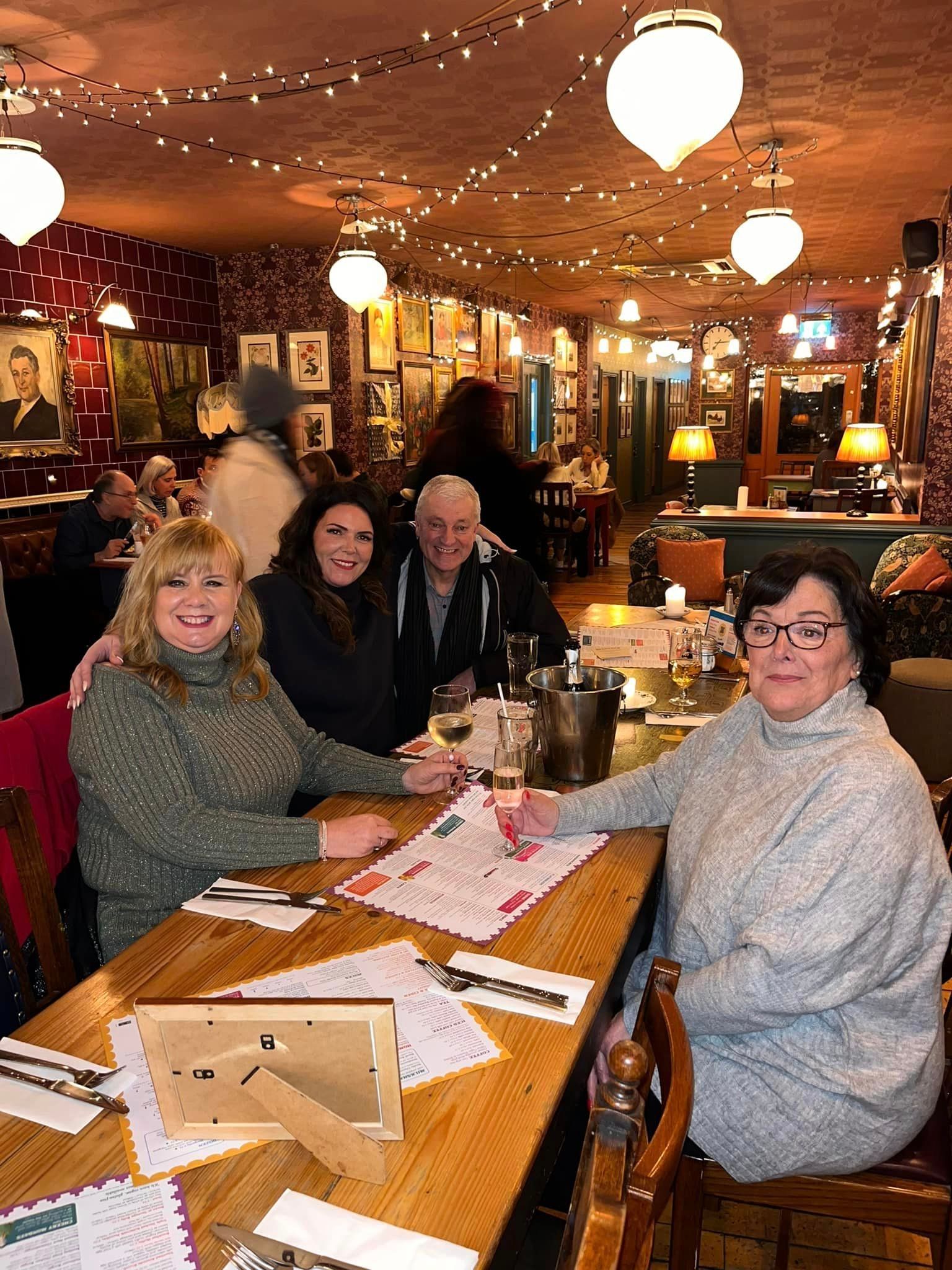 A group of people sits at a wooden table in a dimly lit, decorated restaurant with string lights, enjoying a meal.