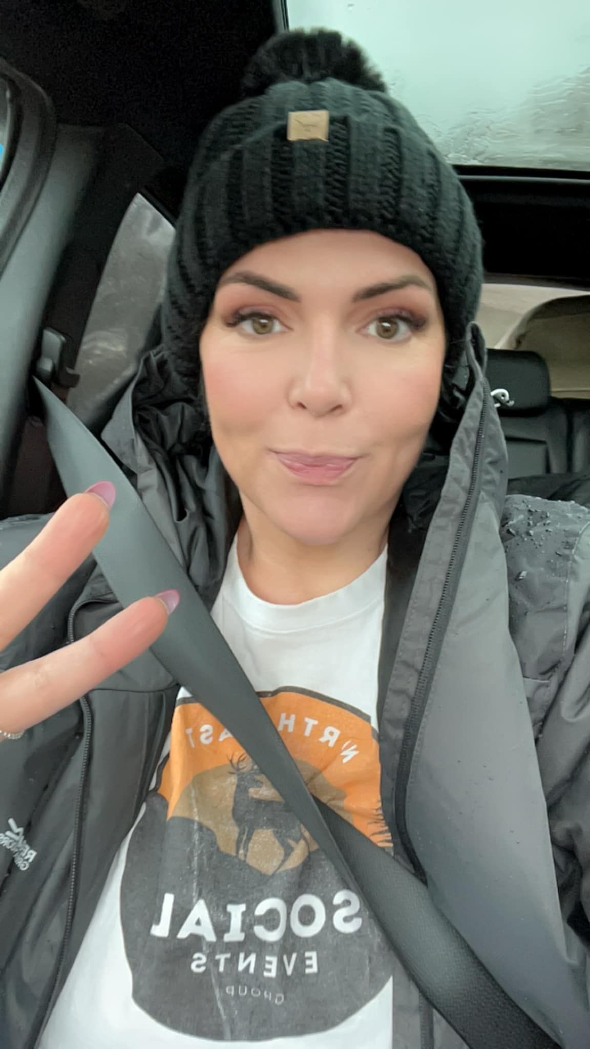 A person wearing a black pom-pom beanie and jacket makes a peace sign while sitting in a car wearing a graphic t-shirt.