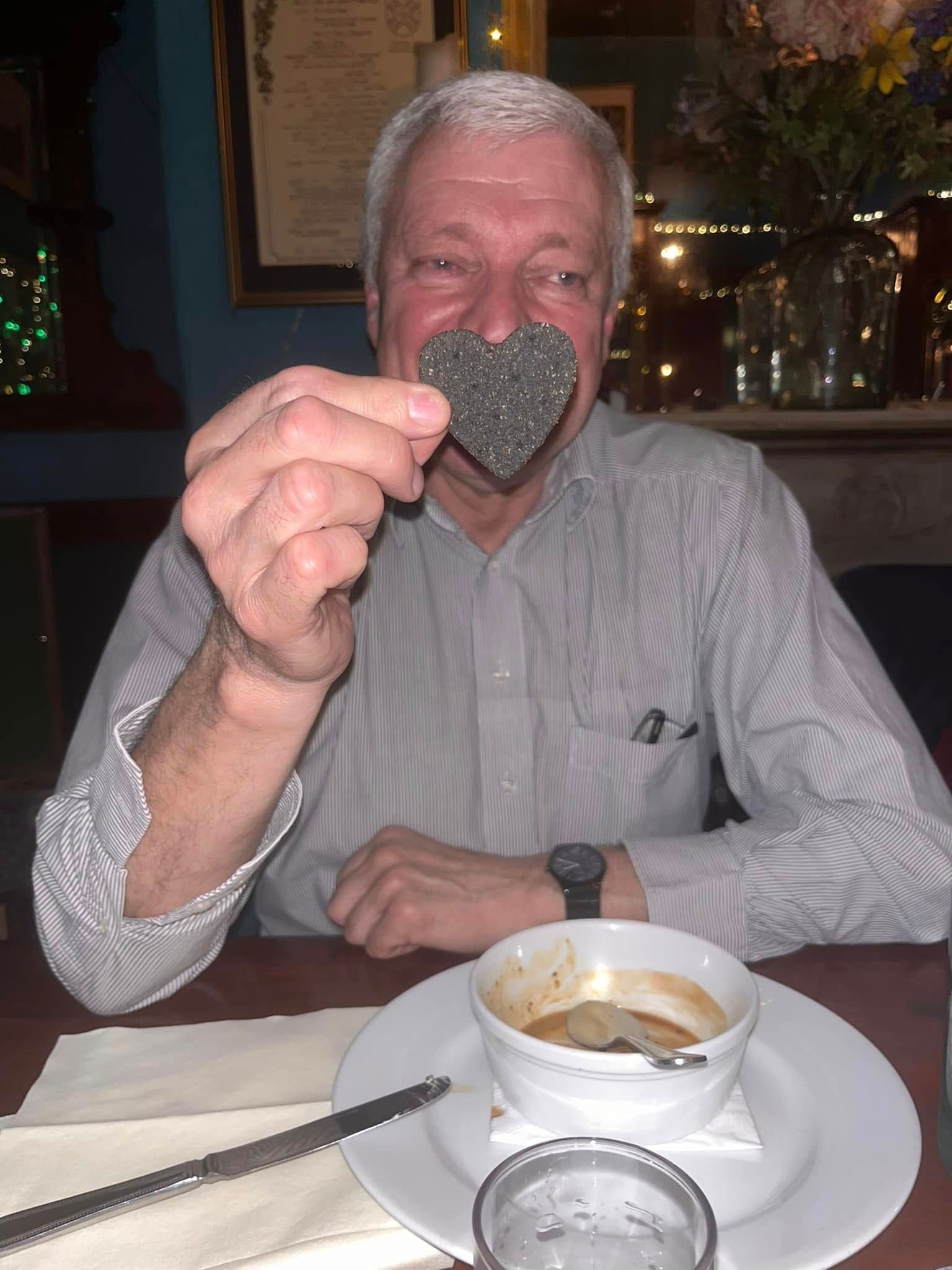 A person sits at a table and holds a heart-shaped, dark textured cracker in front of their nose like a clown nose.