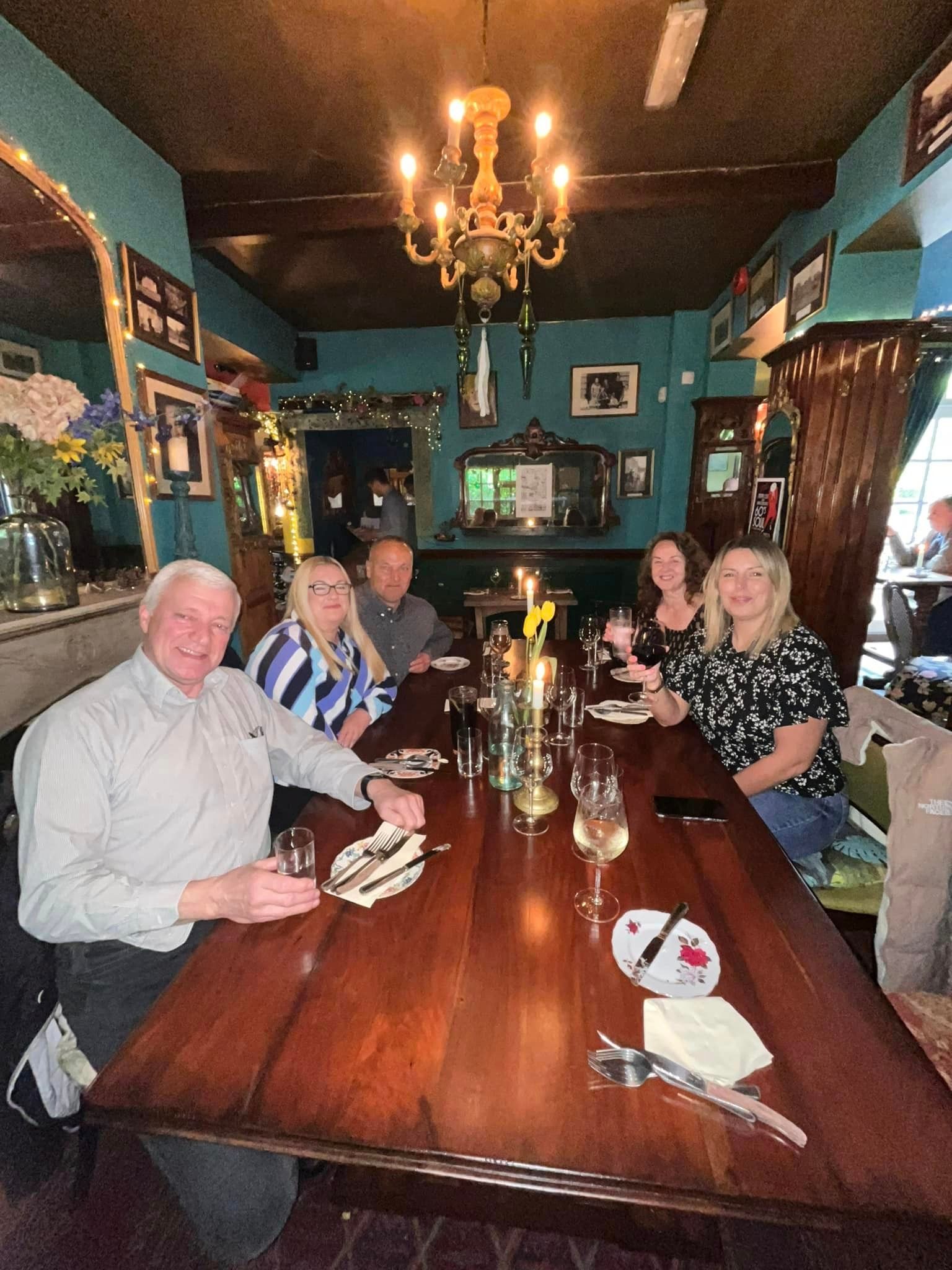 A group of people sit around a long wooden table in a dimly lit, blue-walled restaurant with a chandelier above.