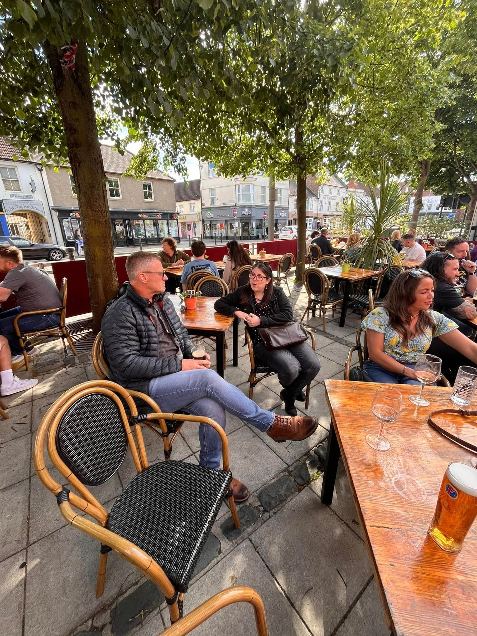 People sit at outdoor tables under trees at a cafe, with a man in a quilted jacket in the foreground.