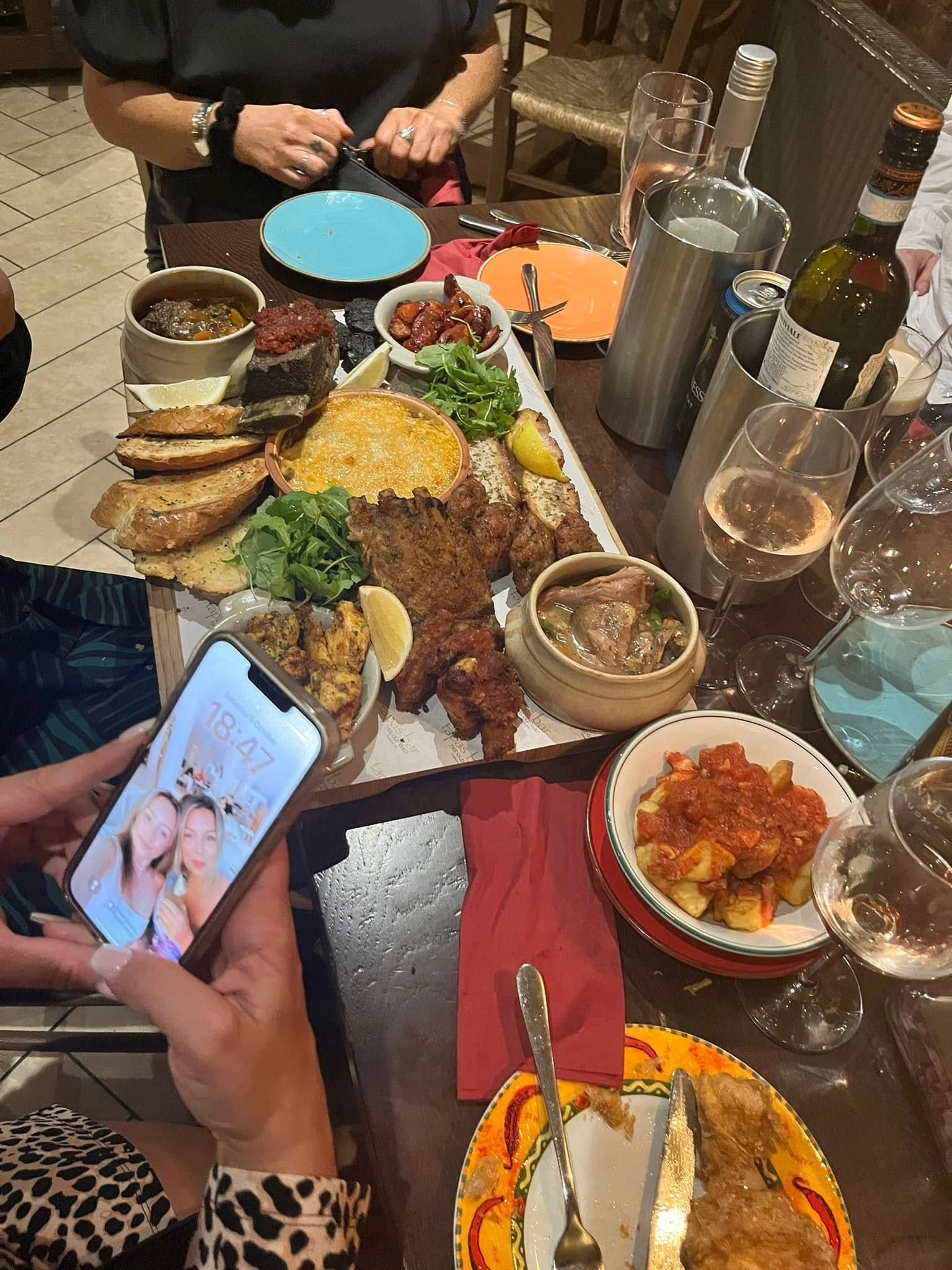 A table filled with shared appetizer platters, glasses of wine, and a person holding a smartphone showing a selfie.