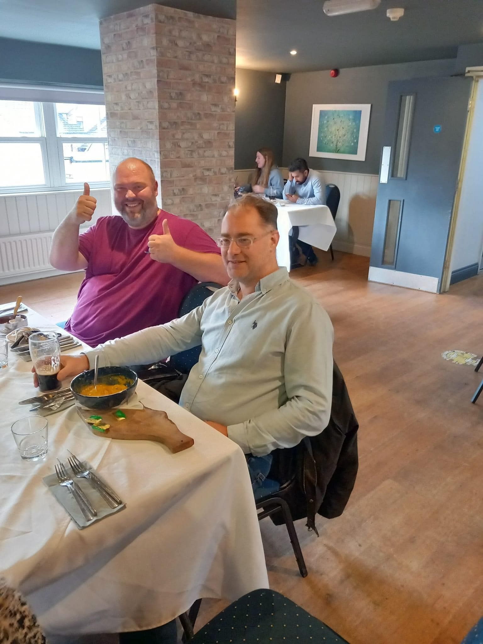 Two people sit at a dining table; one gestures with thumbs up while the other holds a glass in a restaurant setting.