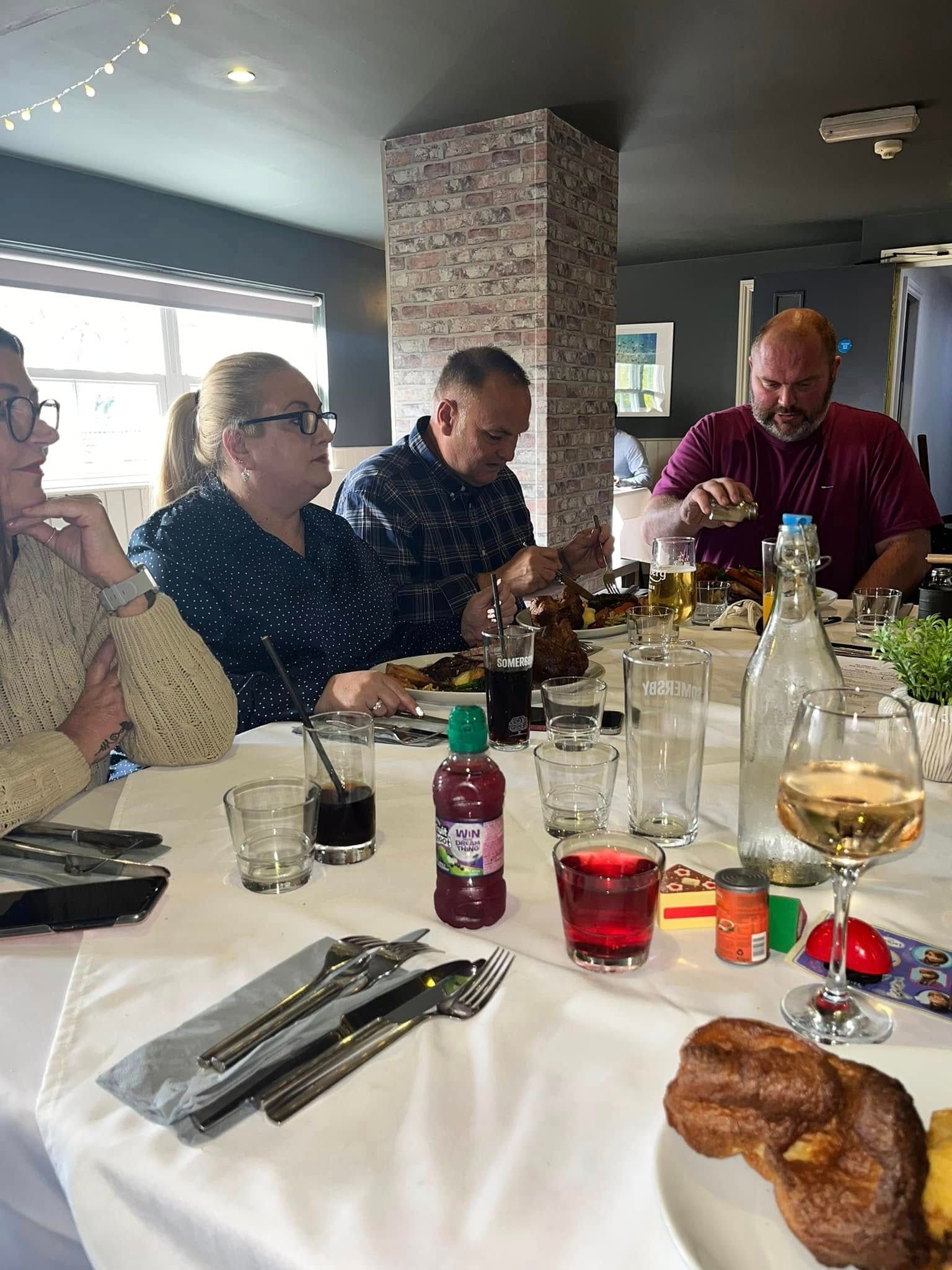 Four people sitting at a table in a restaurant with white tablecloths, glassware, and food items.
