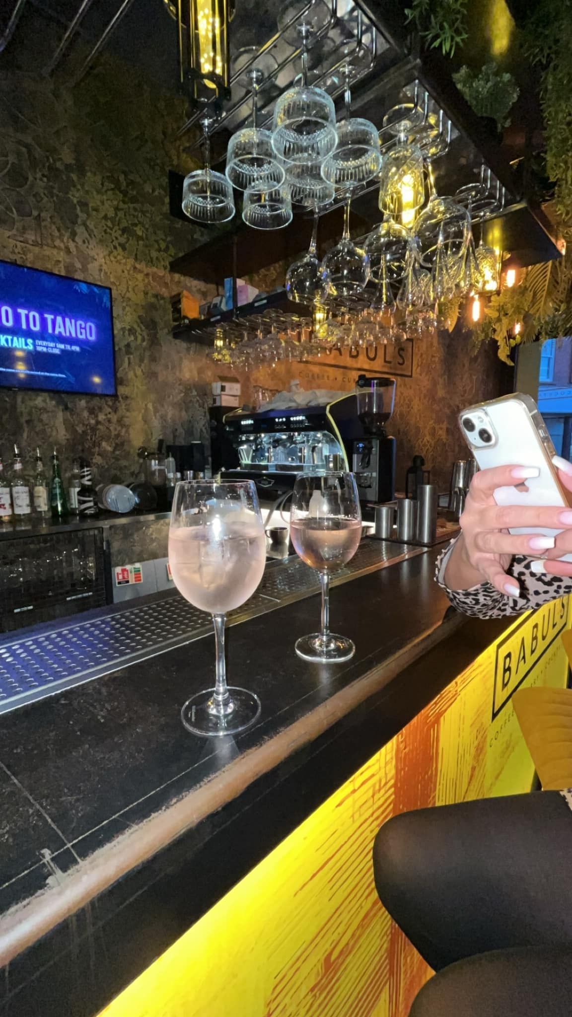 Two glasses of pink cocktails sit on a bar counter, with a hand holding a smartphone in the foreground.