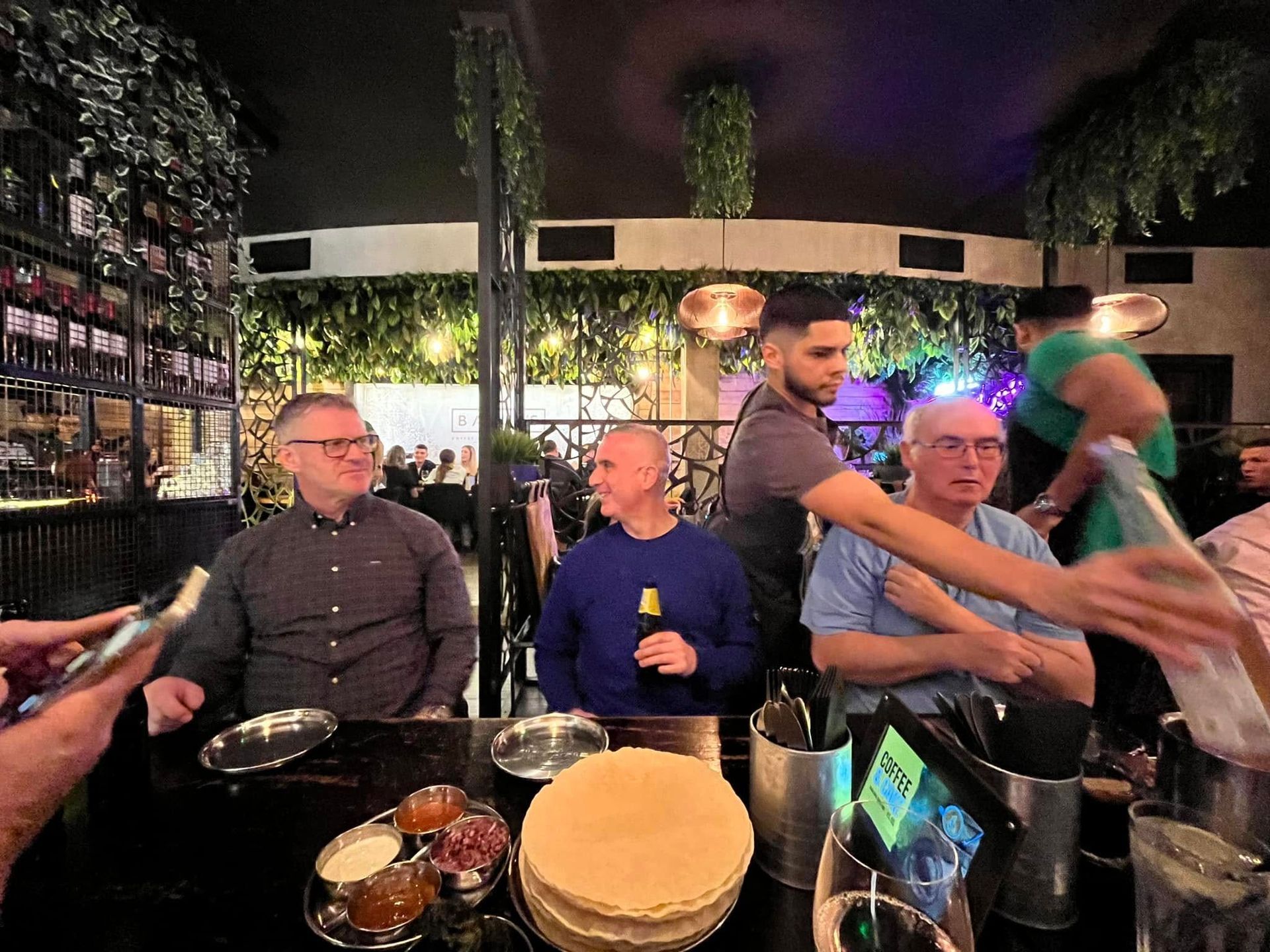 Three people sit at a table in a dimly lit, vine-decorated restaurant as a waiter pours drinks for them.