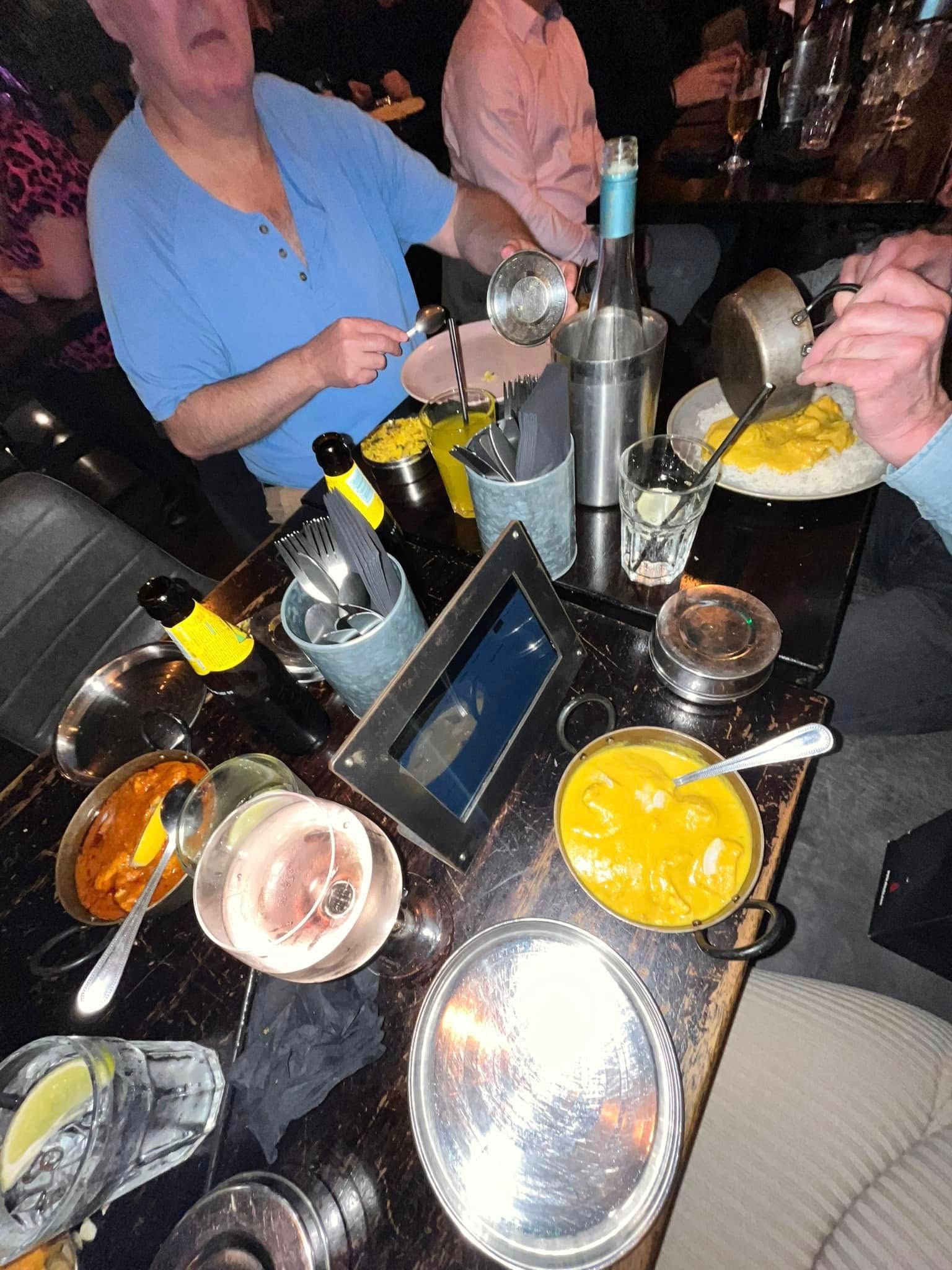 A group of people dining at a table featuring various Indian dishes, metal serving bowls, and drinks in a dim restaurant.