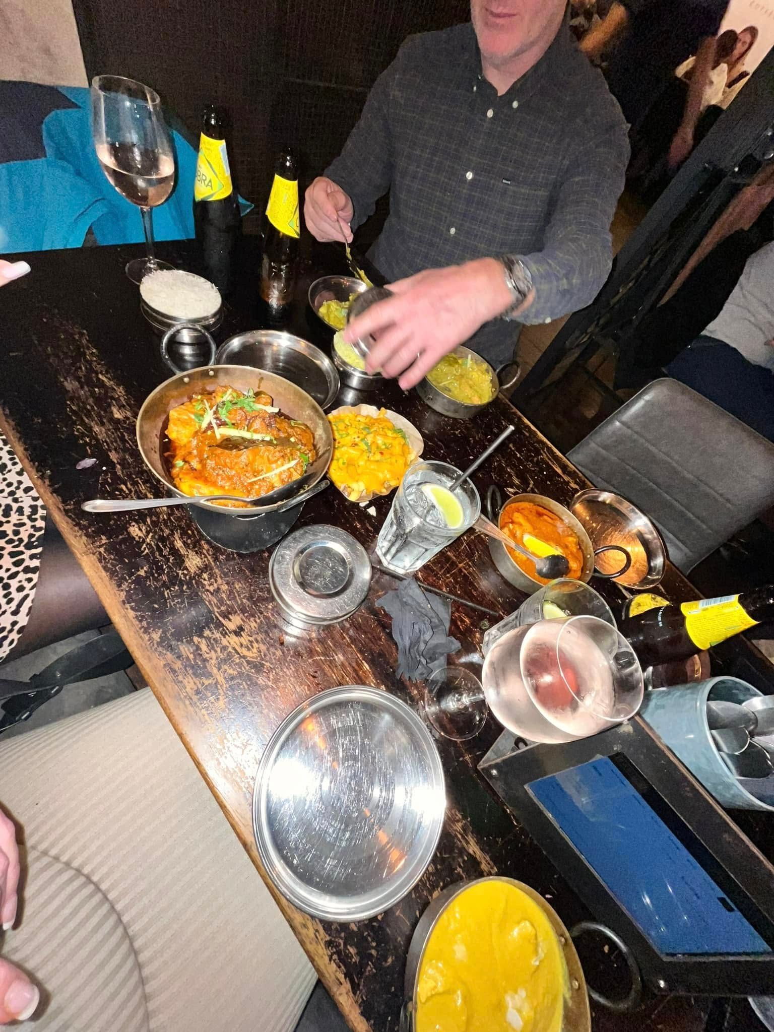 A dining table full of various Indian dishes in metal bowls and beverages, with a person reaching for a serving dish.