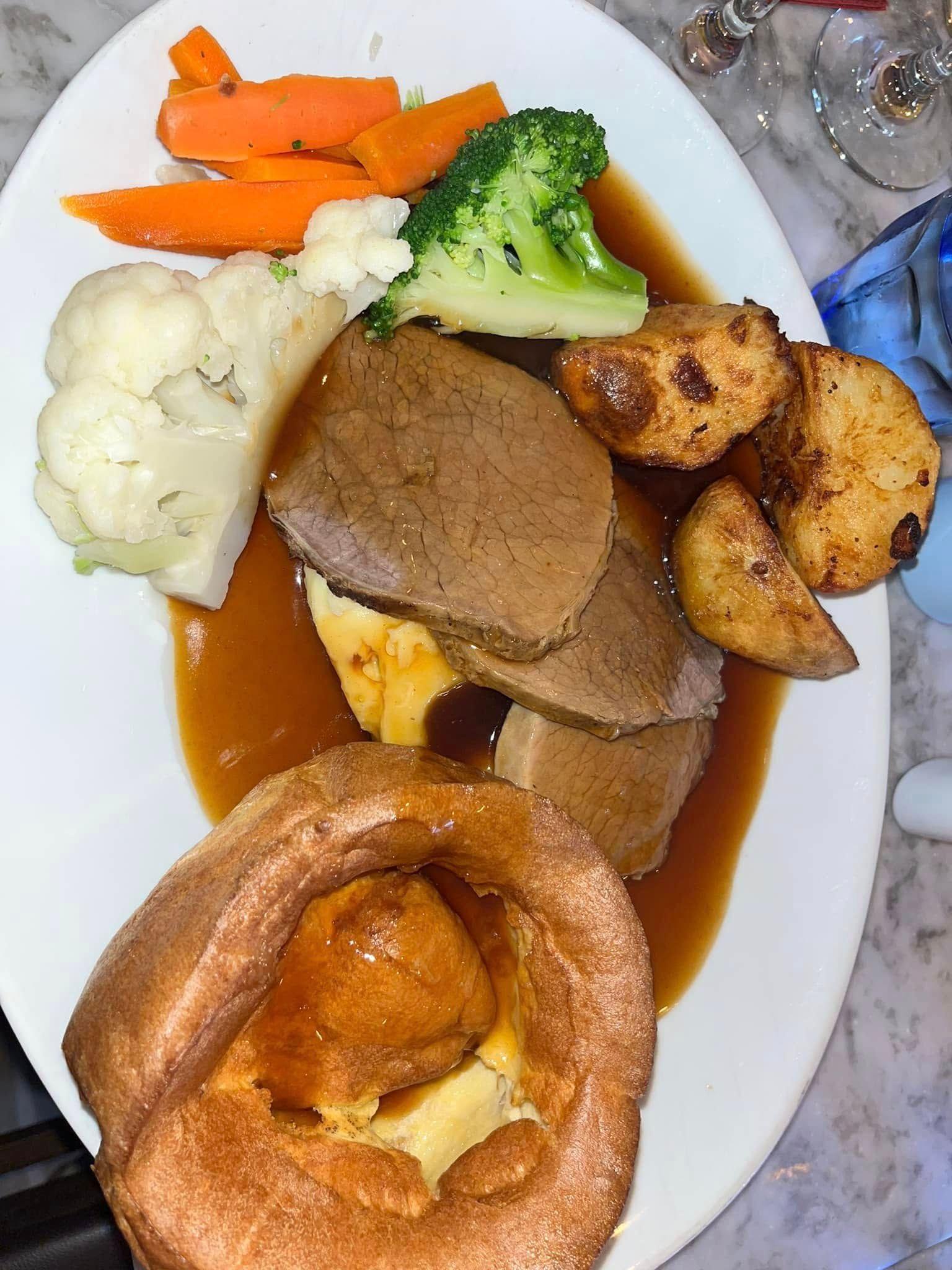 A plate of roast dinner with sliced beef, Yorkshire pudding, roasted potatoes, carrots, broccoli, and cauliflower.