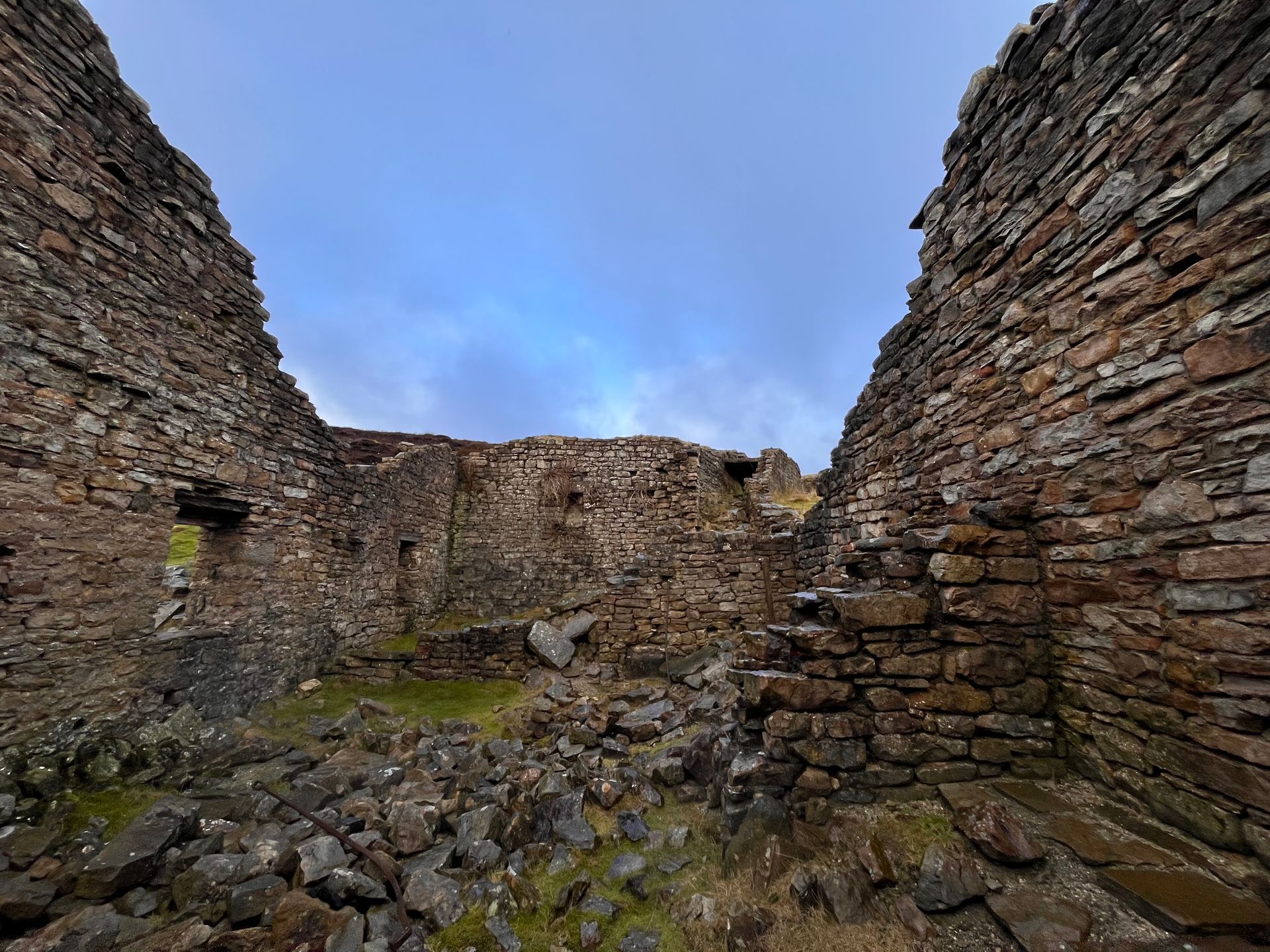 The stone ruins of an ancient building with crumbling walls, a rocky floor, and a backdrop of a cloudy, blue sky.