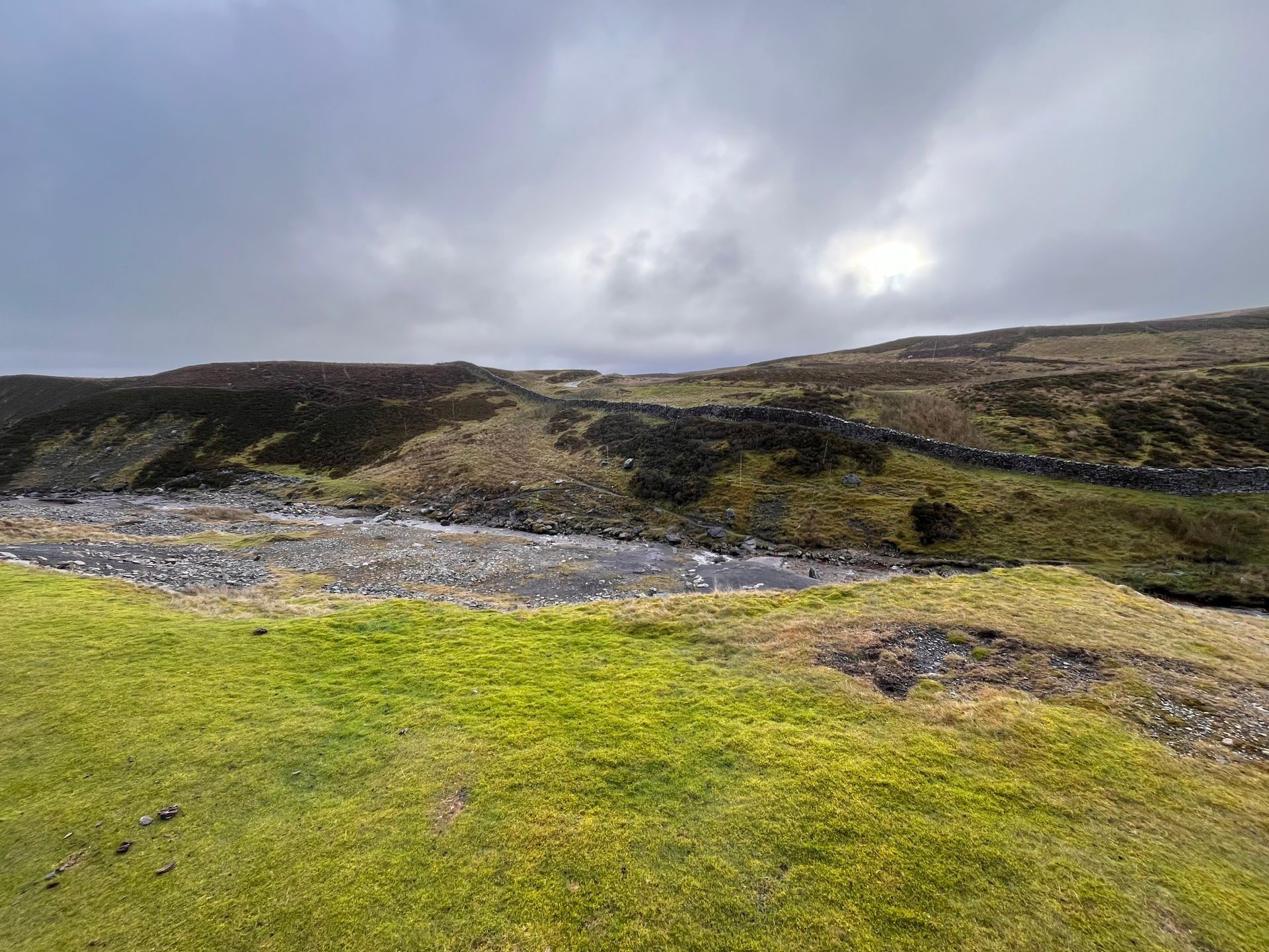 A landscape view of a rocky stream flowing through a grassy moorland under a cloudy sky.