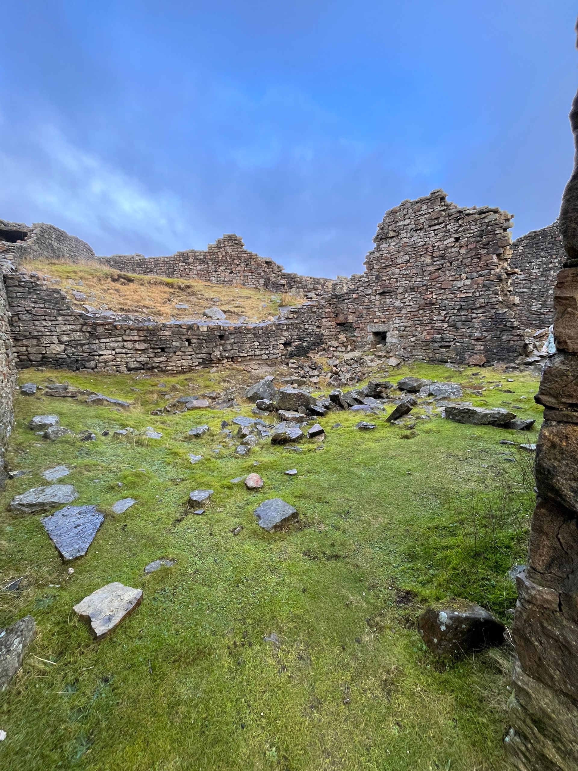 Stone ruins of an ancient building stand on a grassy slope under a cloudy blue sky.