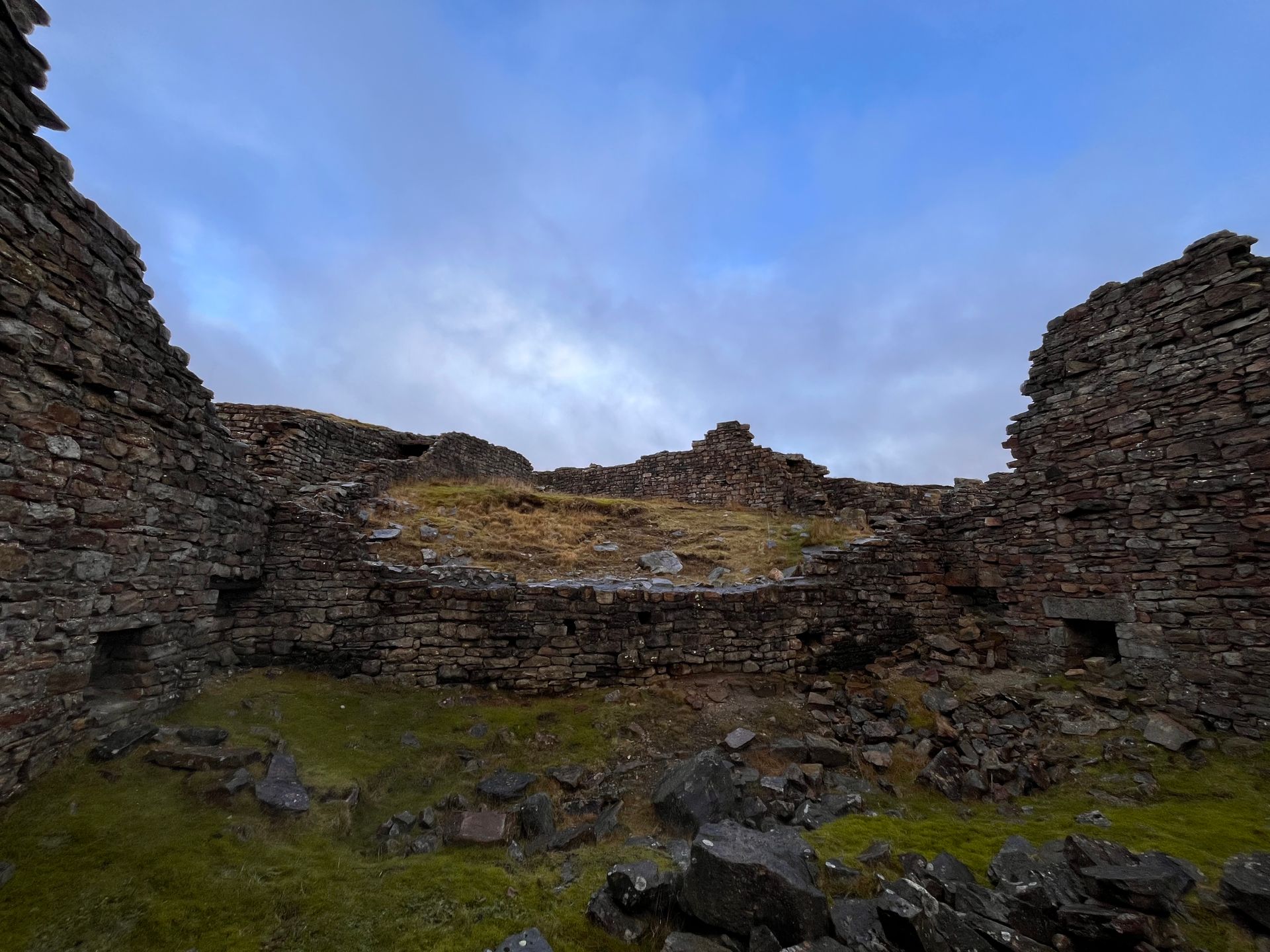 Low-angle view of rugged, mossy stone walls forming the ruins of an ancient circular structure under a cloudy blue sky.