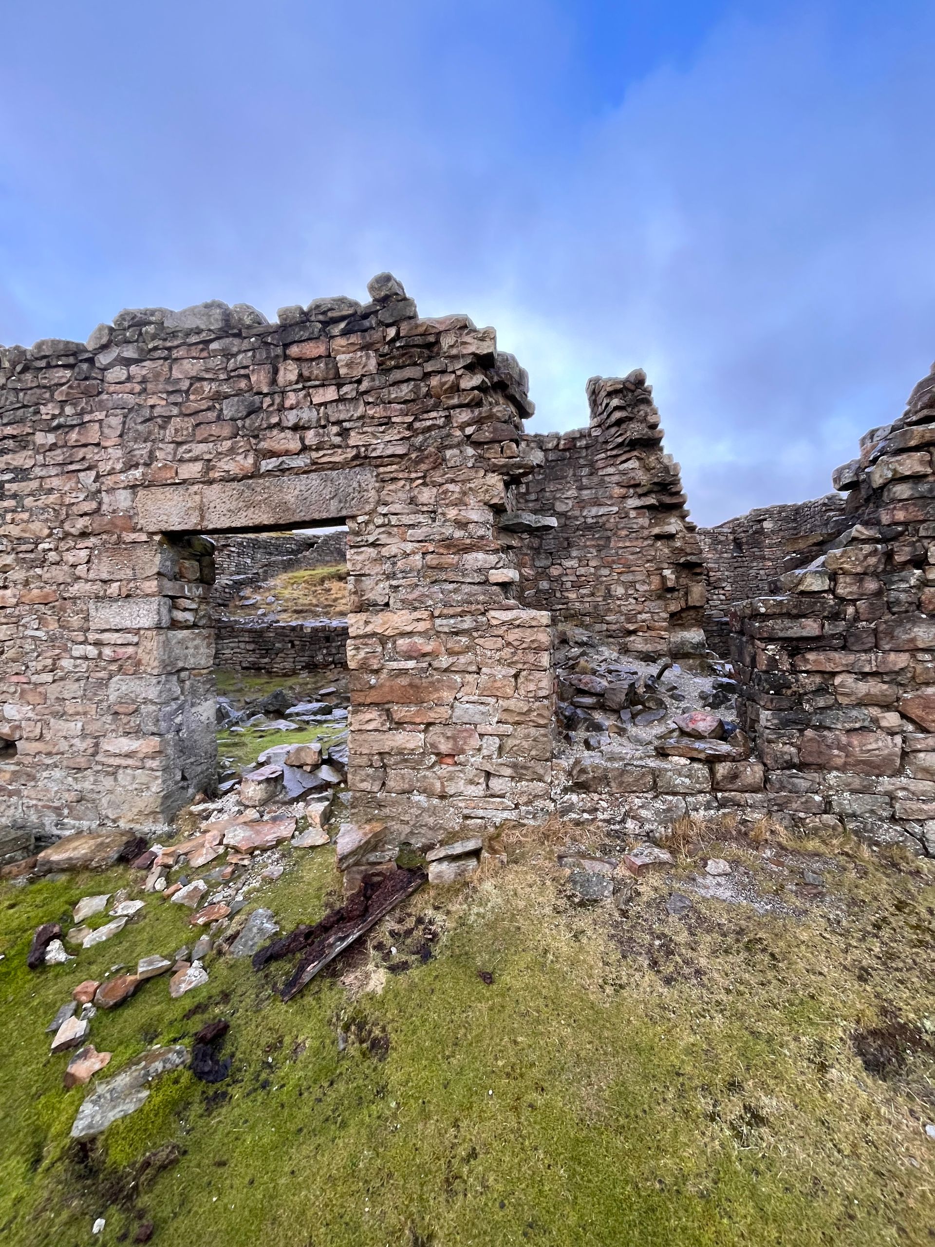 A partially ruined stone building wall with an open doorway, set against a grassy landscape under a blue sky.